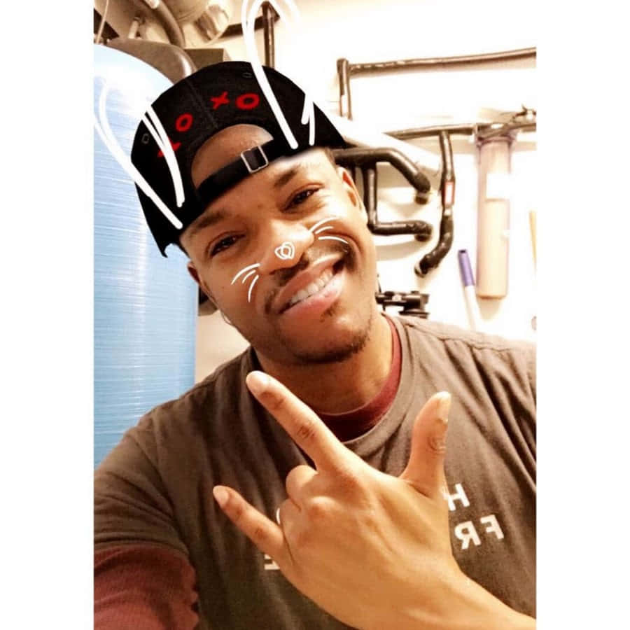Prettyboyfredo 桌布 壁紙 — 一個微笑的男人戴著黑色帽子，上面有紅色XO文字和白色貓鬚濾鏡，在室內做出搖滾手勢。
