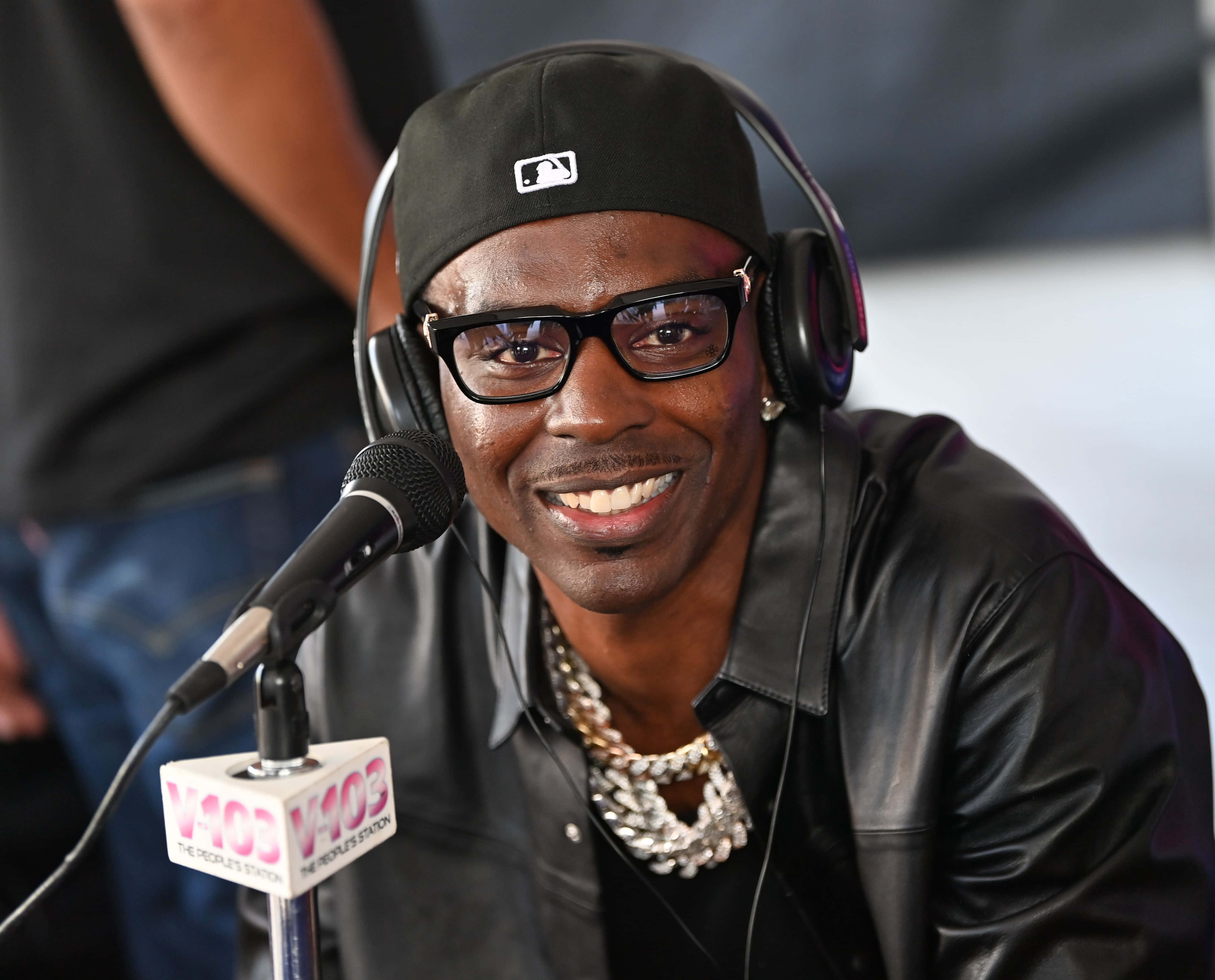 Young Dolph 桌布 壁紙 — 一個微笑的男人戴著黑色帽子和皮夾克，戴著耳機，對著V103電台麥克風說話。