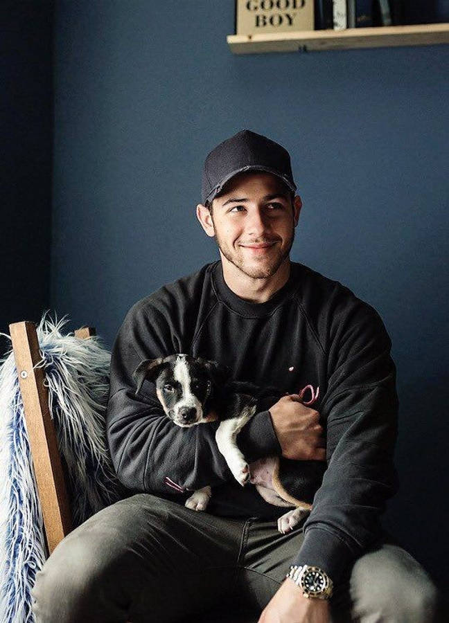 Jonas Brothers 桌布 壁紙 — 一個微笑的男人戴著黑色鴨舌帽和運動衫，室內坐著，抱著一隻黑白小狗，背景是深藍色牆壁。