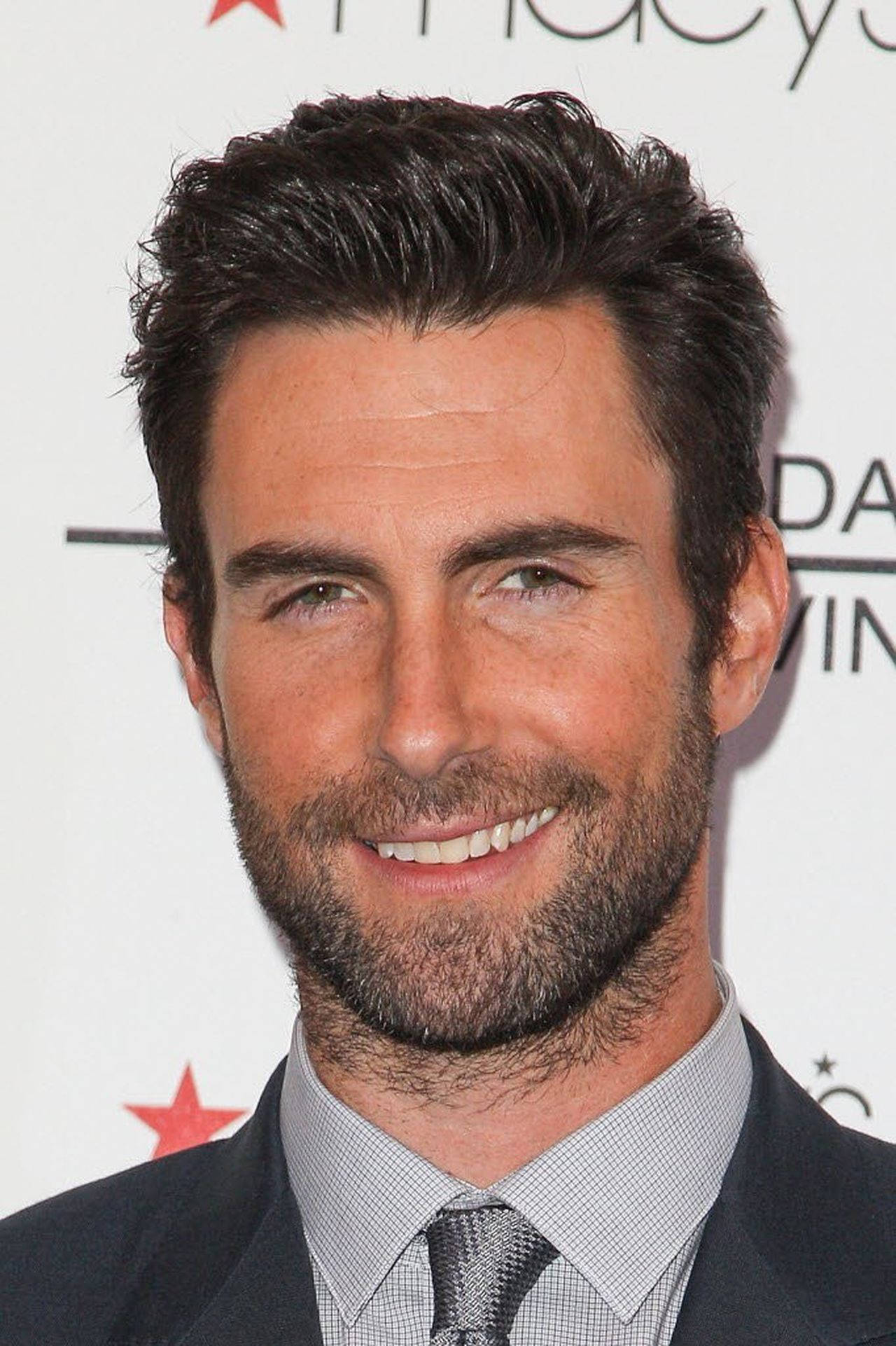 Adam Levine 桌布 壁紙 — 一個微笑的男人特寫肖像，深色頭髮和鬍渣，穿著西裝和領帶，背景為品牌背景。