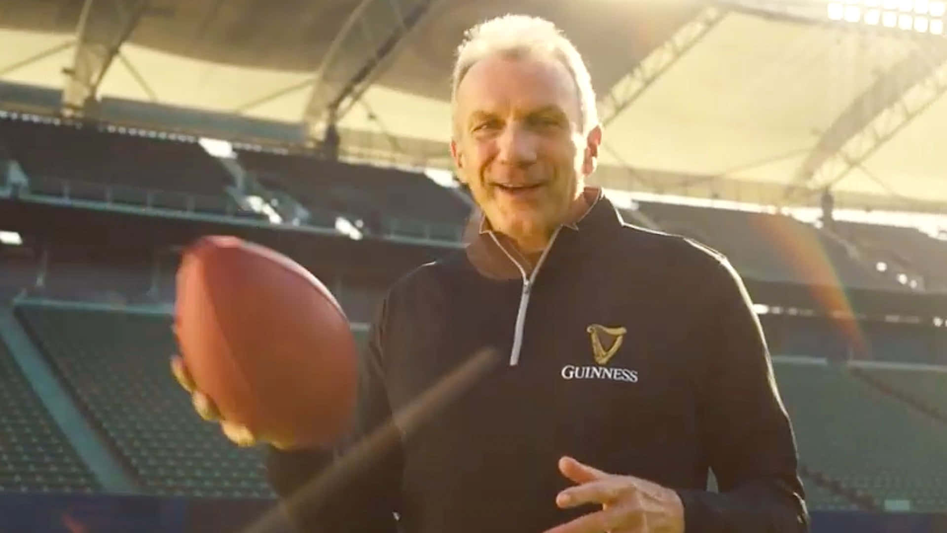 Joe Montana 桌布 壁紙 — 一個微笑的男人穿著Guinness外套，在陽光明媚的體育場裡扔足球，身後是空座位。