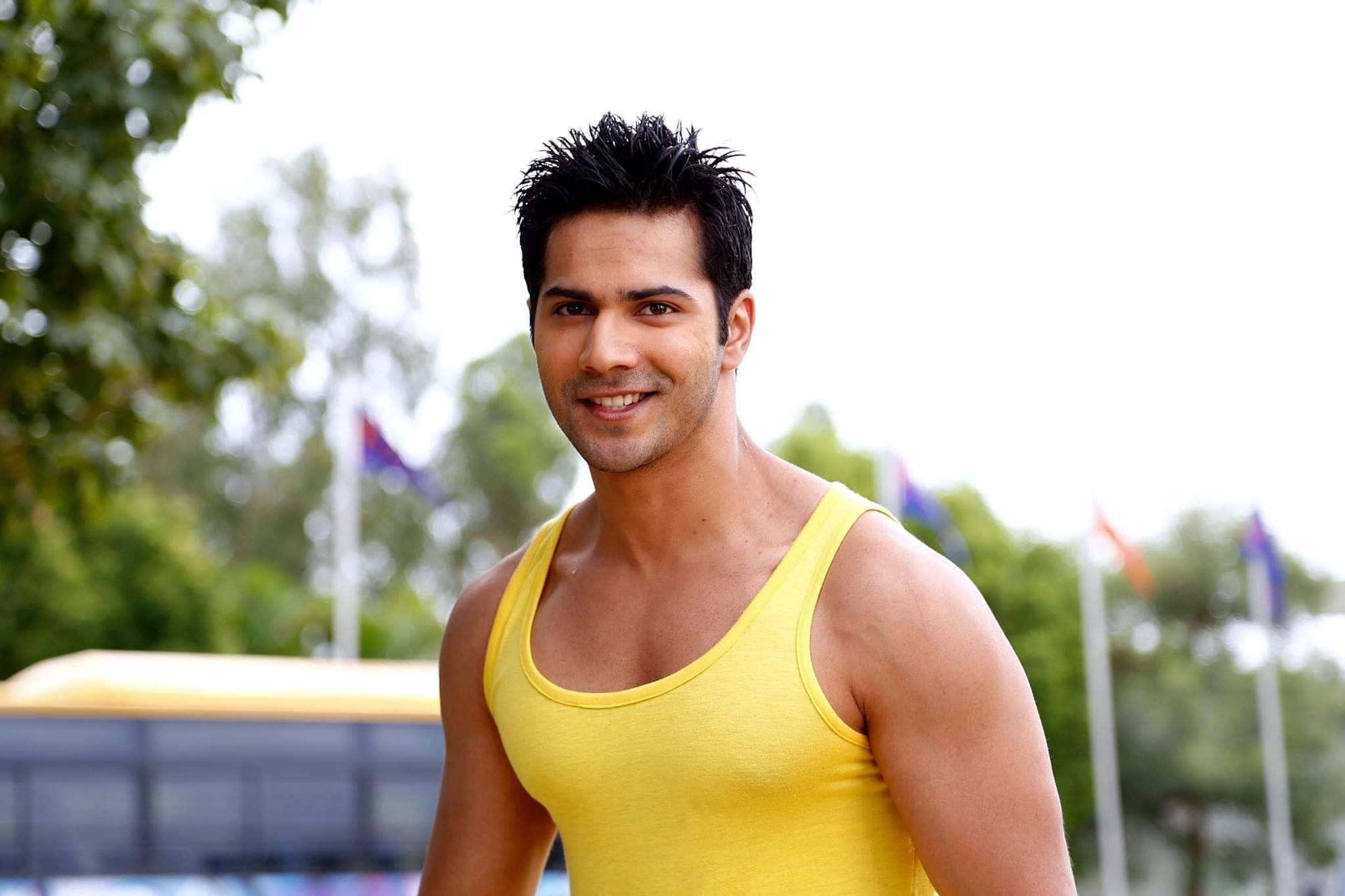 Varun Dhawan 桌布 壁紙 — 一個微笑的男人穿著亮黃色背心，站在戶外，背景有樹木和旗幟。