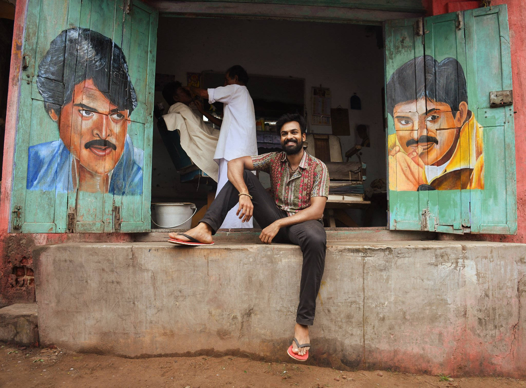 Indian 桌布 壁紙 — 一個微笑的男人穿著人字拖坐在商店前的水泥階梯上，商店有繪有大型男性肖像的油漆木製百葉窗。