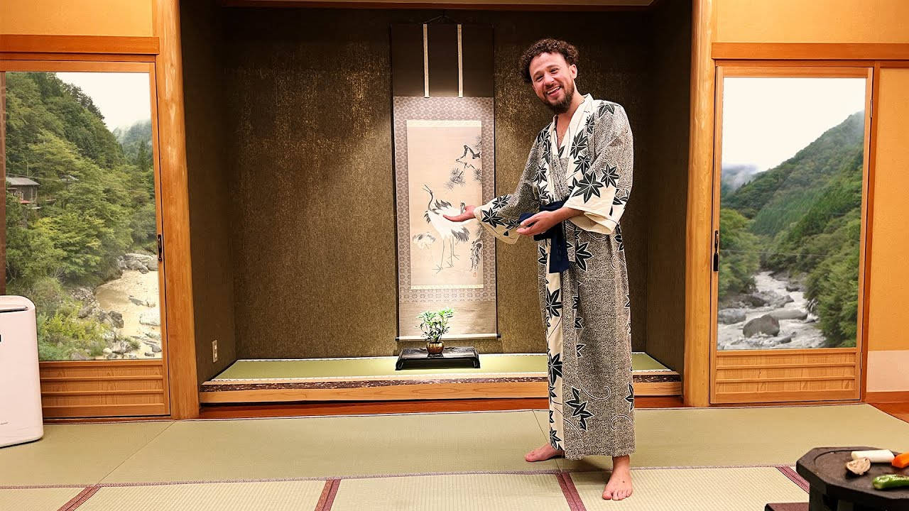 灰色 桌布 壁紙 — 一個微笑的男人穿著圖案浴衣，赤腳站在傳統的日式房間裡，房間裡有榻榻米和推拉門，門外是鬱鬱蔥蔥的山河景色。