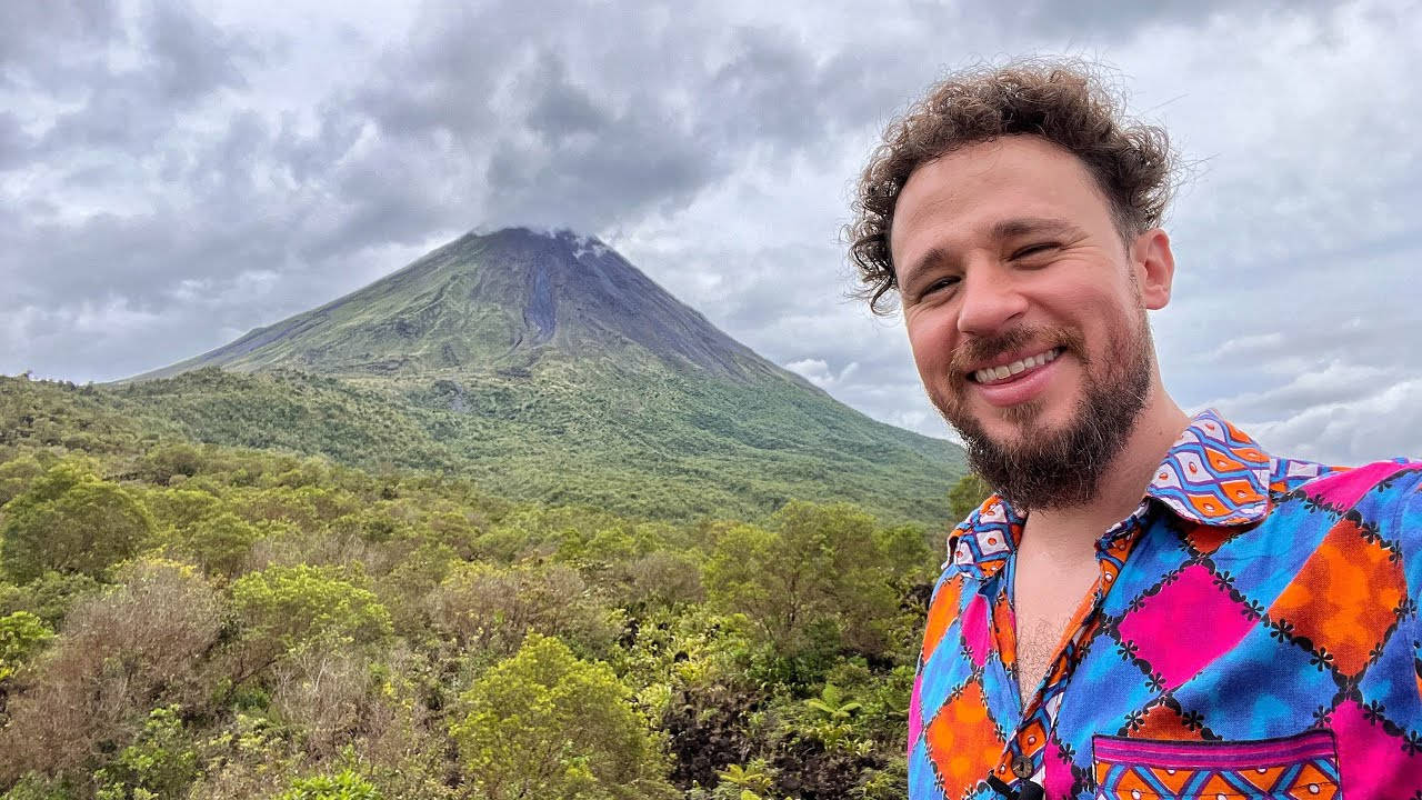 Volcano 桌布 壁紙 — 一個微笑的男人穿著彩色圖案襯衫，站在多雲天空下茂密的綠色火山前。