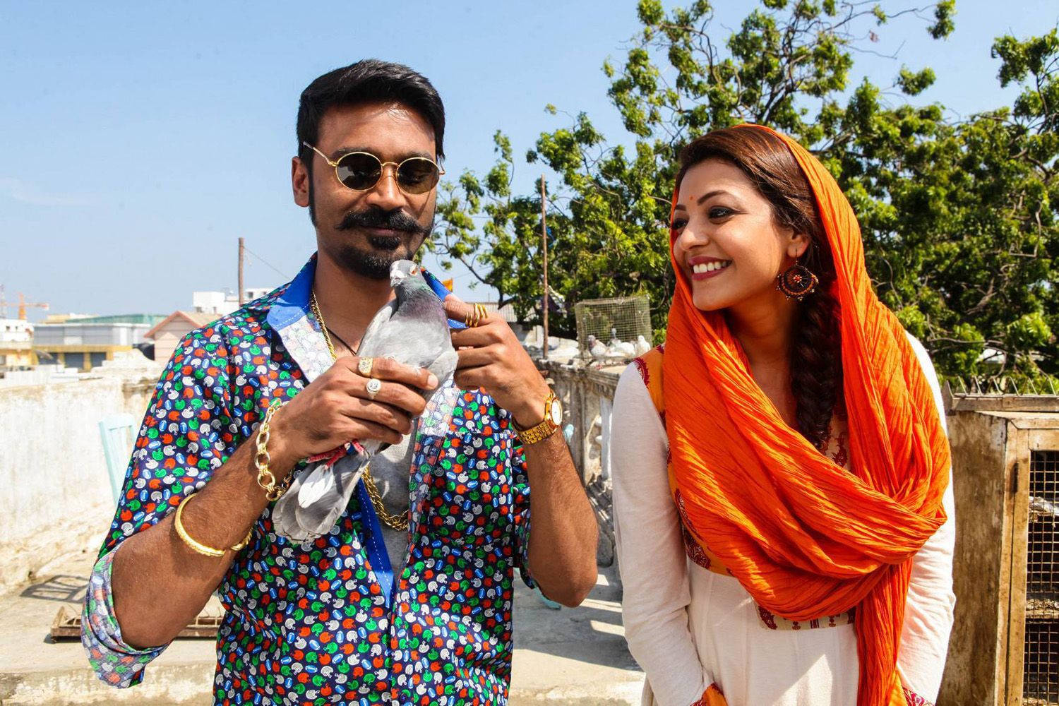 Bollywood HD 桌布 壁紙 — 一個微笑的男人穿著彩色襯衫，在明亮的藍天下，站在一位戴著橙色圍巾的女人旁邊，抱著一隻鴿子。
