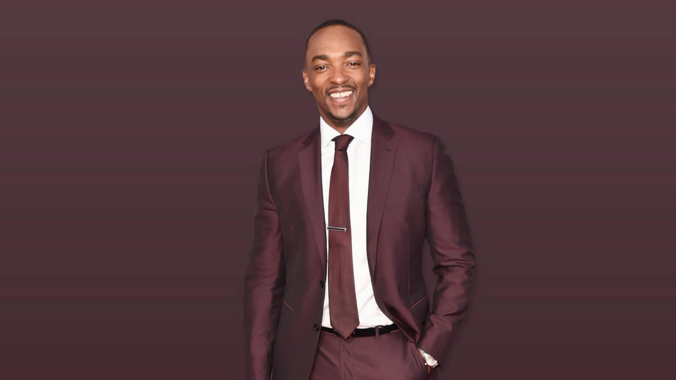 Anthony Mackie 桌布 壁紙 — 一個微笑的男人穿著栗色西裝，自信地站在紫色漸變背景前。