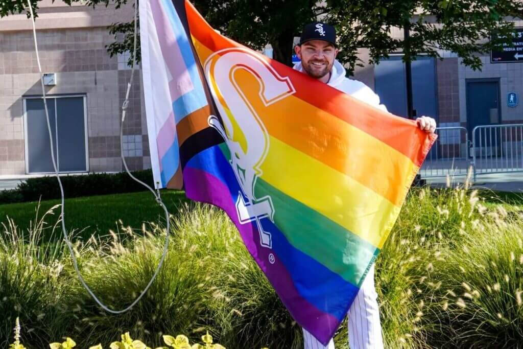 Chicago White Sox 桌布 壁紙 — 一個微笑的男人穿著棒球制服，在陽光明媚的日子裡戶外舉著帶有白色「S」標誌的彩虹旗。