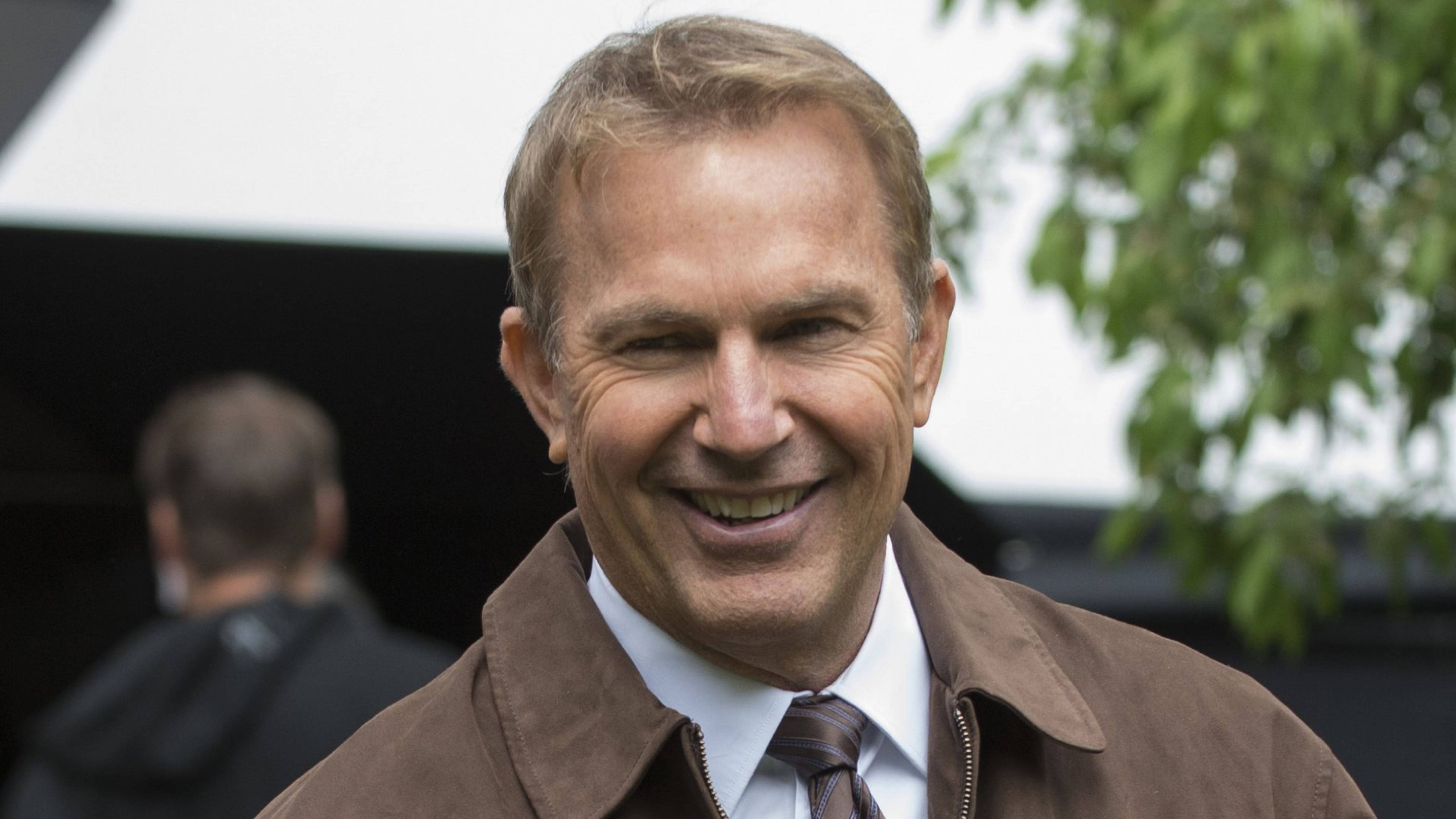 Kevin Costner 桌布 壁紙 — 一個微笑的男人穿著棕色外套和領帶，站在戶外，身後有綠色樹葉。