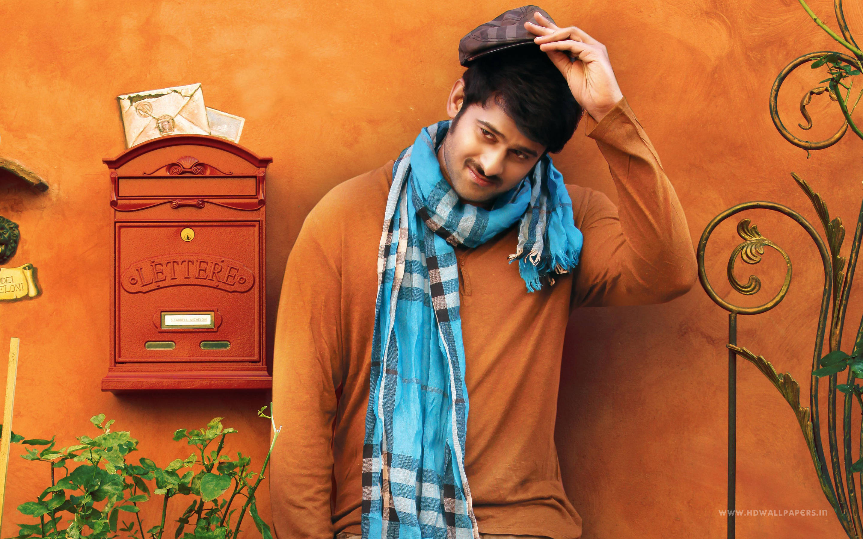 Prabhas HD 桌布 壁紙 — 一個微笑的男人穿著棕色毛衣和藍色格紋圍巾，在溫暖的橙色牆壁旁調整他的帽子，旁邊有一個紅色郵筒。