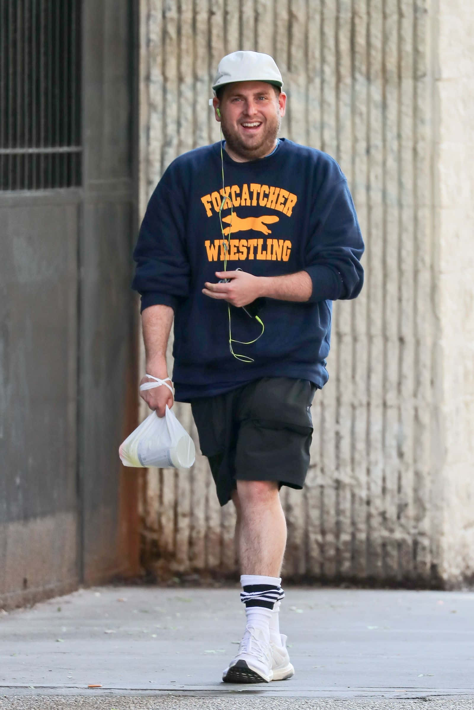 jonah hill 桌布 壁紙 — 一個微笑的男人穿著海軍藍色 'Foxcatcher Wrestling' 運動衫和白色帽子，拿著一個塑膠袋走在混凝土小巷中。