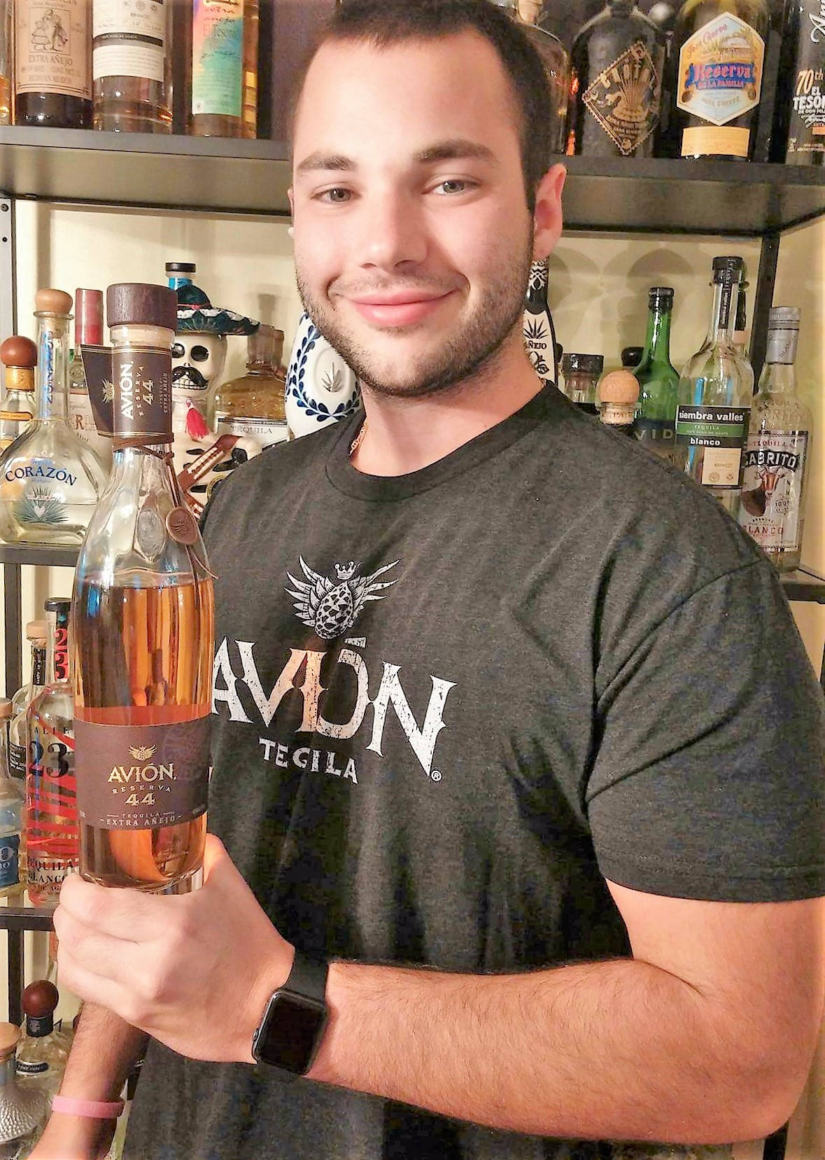 Tequila 桌布 壁紙 — 一個微笑的男人穿著深色T恤，在擺滿酒品的酒吧架前拿著一瓶Avion Tequila。