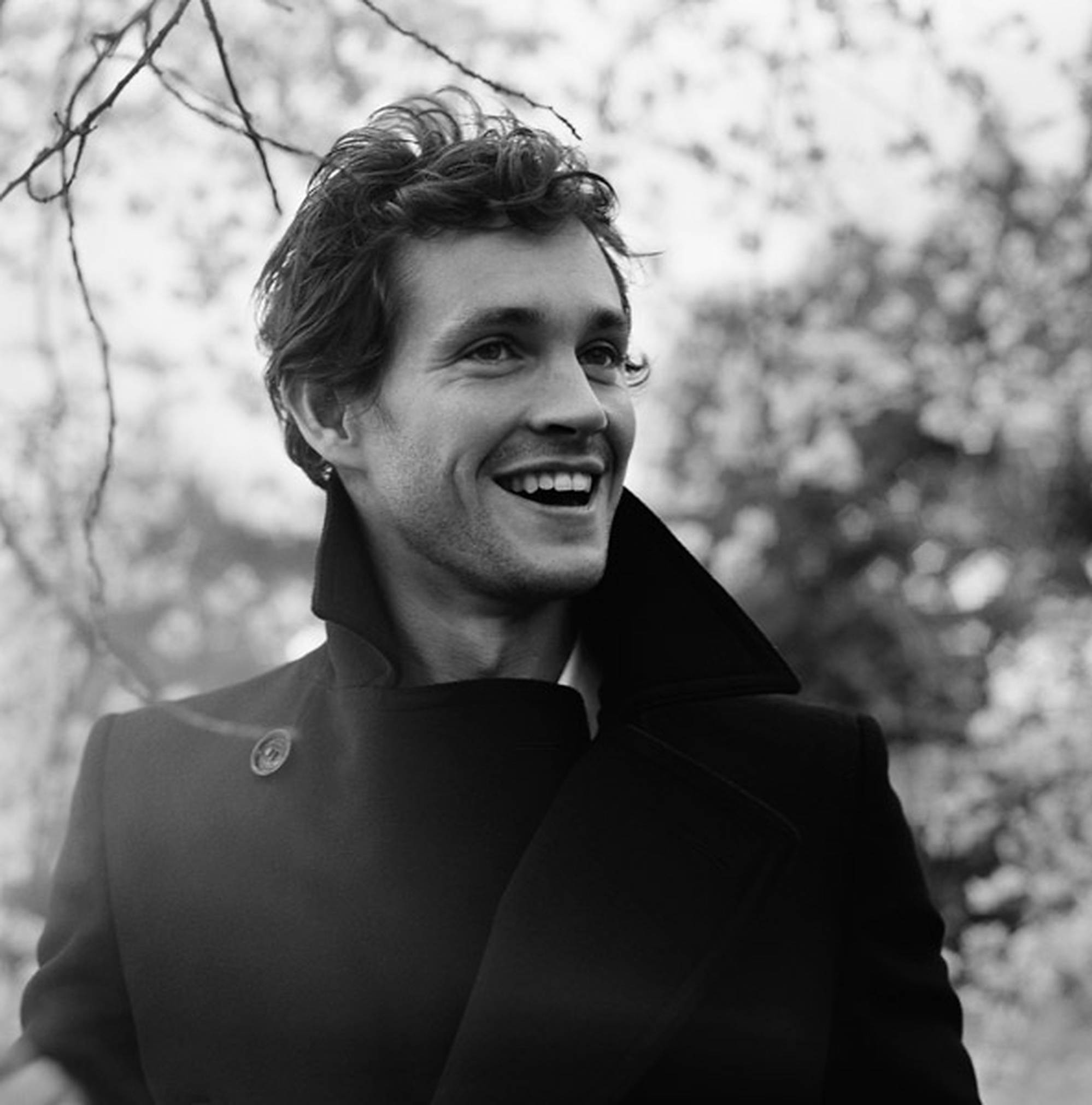 Hugh Dancy 桌布 壁紙 — 一個微笑的男人穿著深色外套站在戶外，身後有模糊樹枝的黑白肖像。