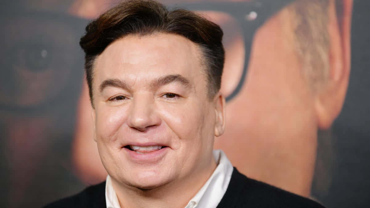 Mike Myers 桌布 壁紙 — 一個微笑的男人穿著深色毛衣，背景是模糊的宣傳背景的近距離照片。