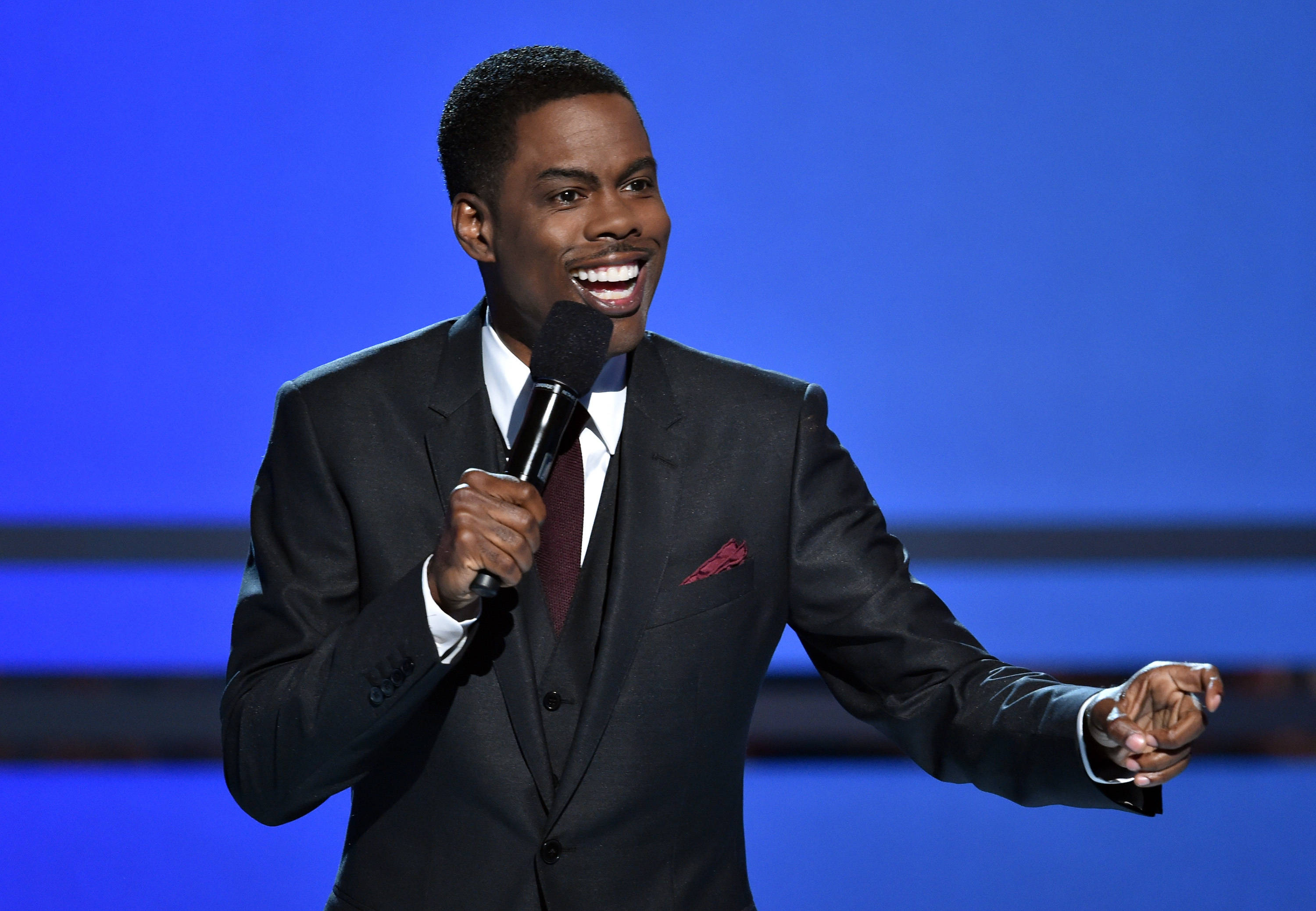 Chris Rock 桌布 壁紙 — 一個微笑的男人穿著深色西裝，在純藍色舞台背景下拿著麥克風。