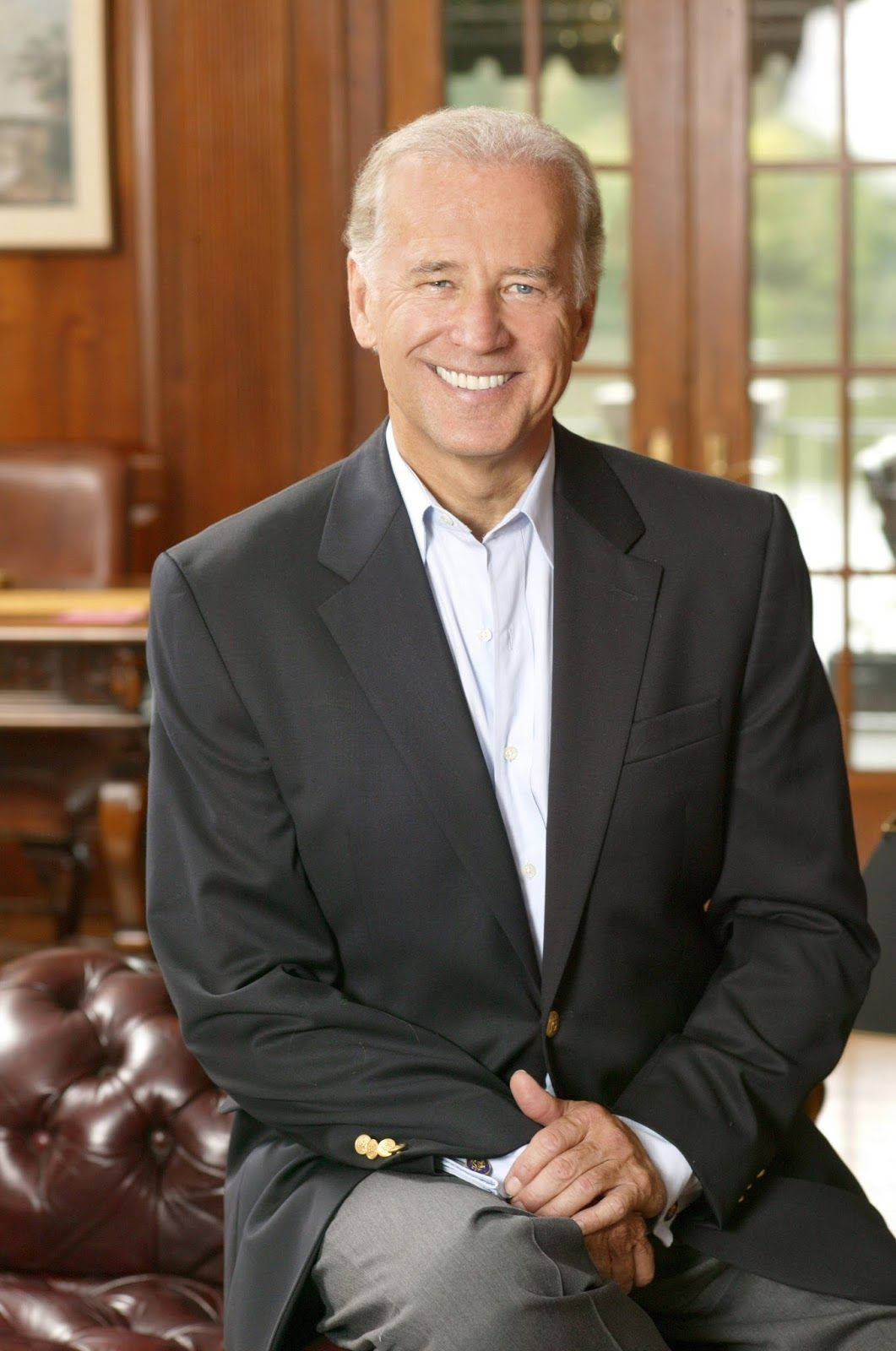 Joe Biden 桌布 壁紙 — 一個微笑的男人穿著深色西裝，自信地坐在一個有木鑲板房間的紅色皮革簇絨椅上，旁邊有大窗戶。