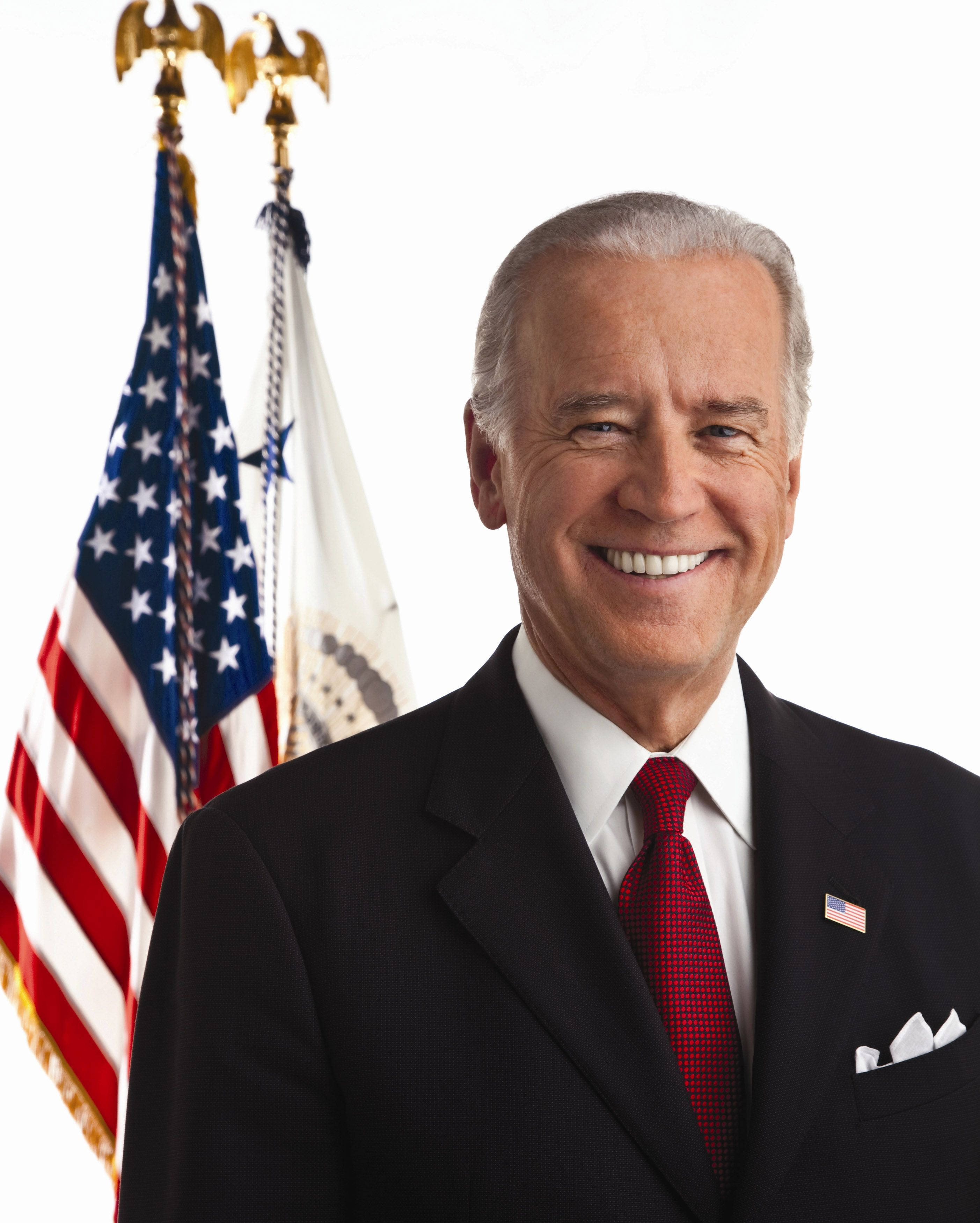 Biden 桌布 壁紙 — 一個微笑的男人穿著深色西裝和紅色領帶，站在兩面旗幟前，背景為白色。