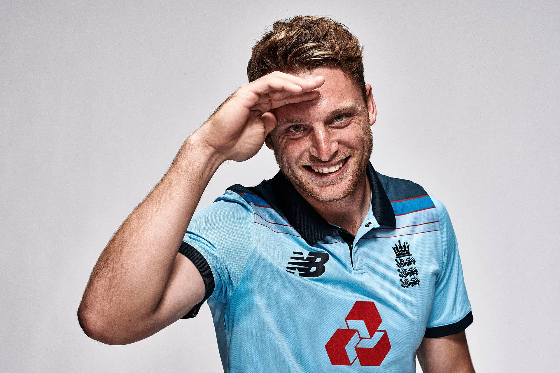 jos buttler 桌布 壁紙 — 一個微笑的男人穿著淺藍色英格蘭板球衫，用手遮住眼睛，背景是純灰色。