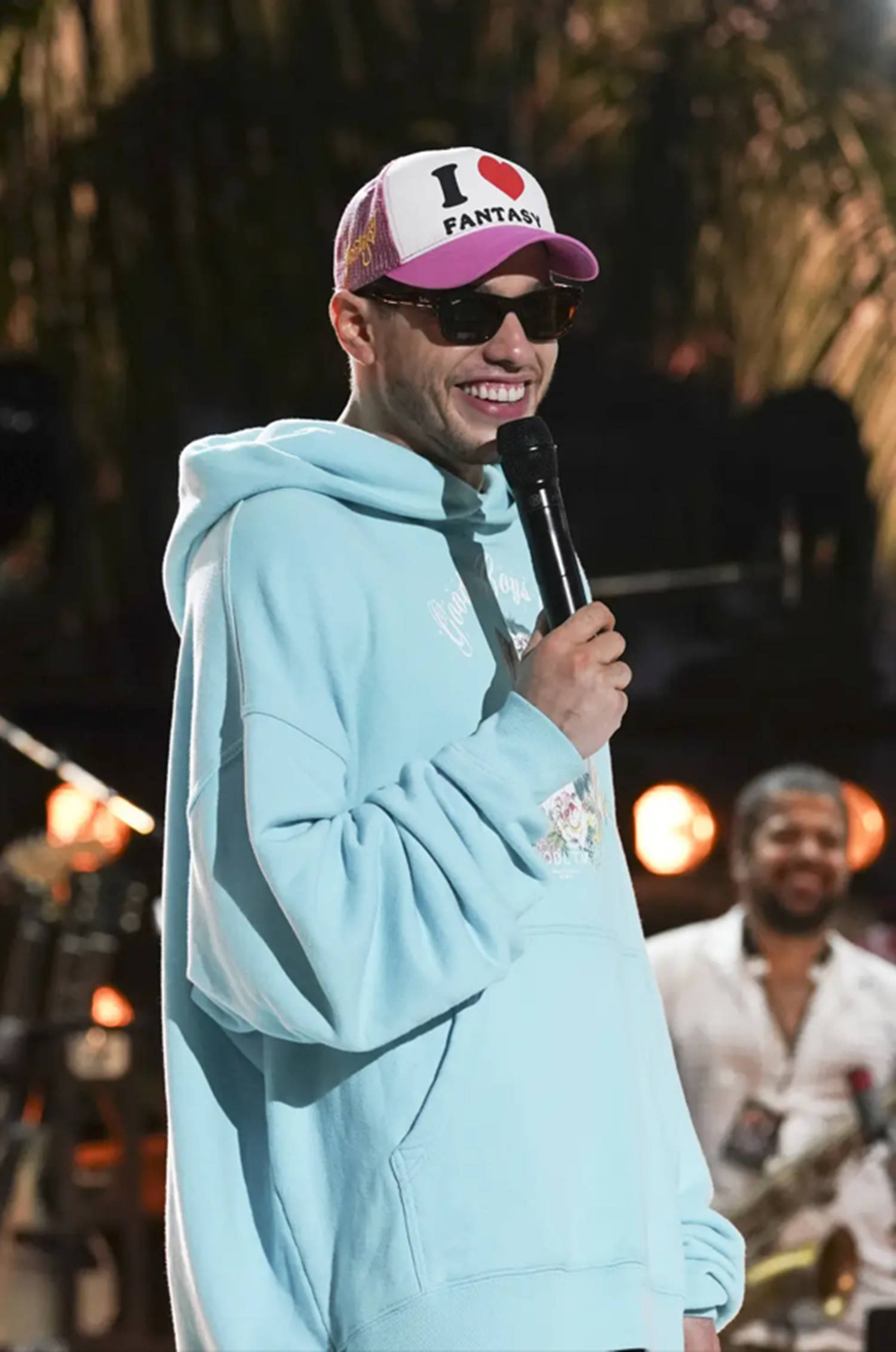 Celebrity 桌布 壁紙 — 一個微笑的男人穿著淺藍色連帽衫和粉紅色帽子，在溫暖燈光的舞台上拿著麥克風，身後有模糊的音樂家。