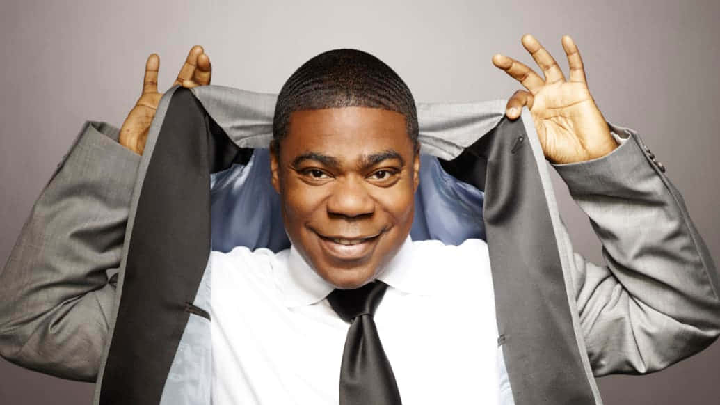 Tracy Morgan 桌布 壁紙 — 一個微笑的男人穿著灰色西裝，掀起外套露出藍色襯衫和黑色領帶，背景是純灰色。