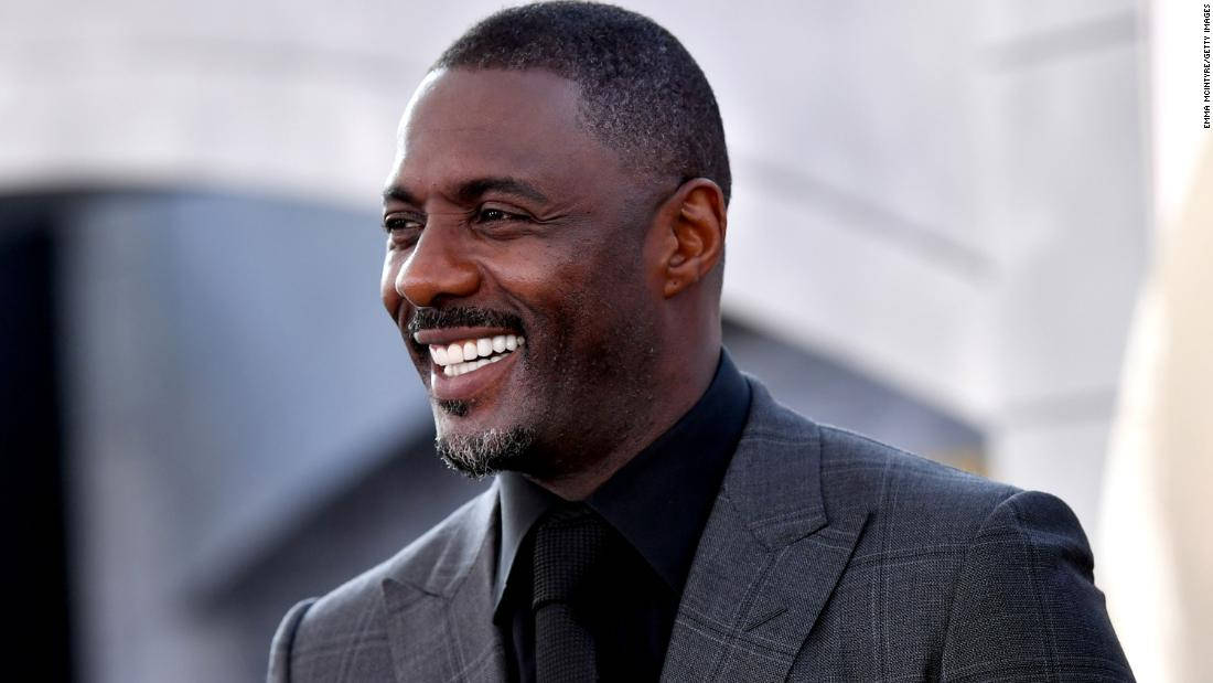 idris elba 桌布 壁紙 — 一個微笑的男人穿著灰色西裝和黑色領帶，站在模糊的現代建築背景前。