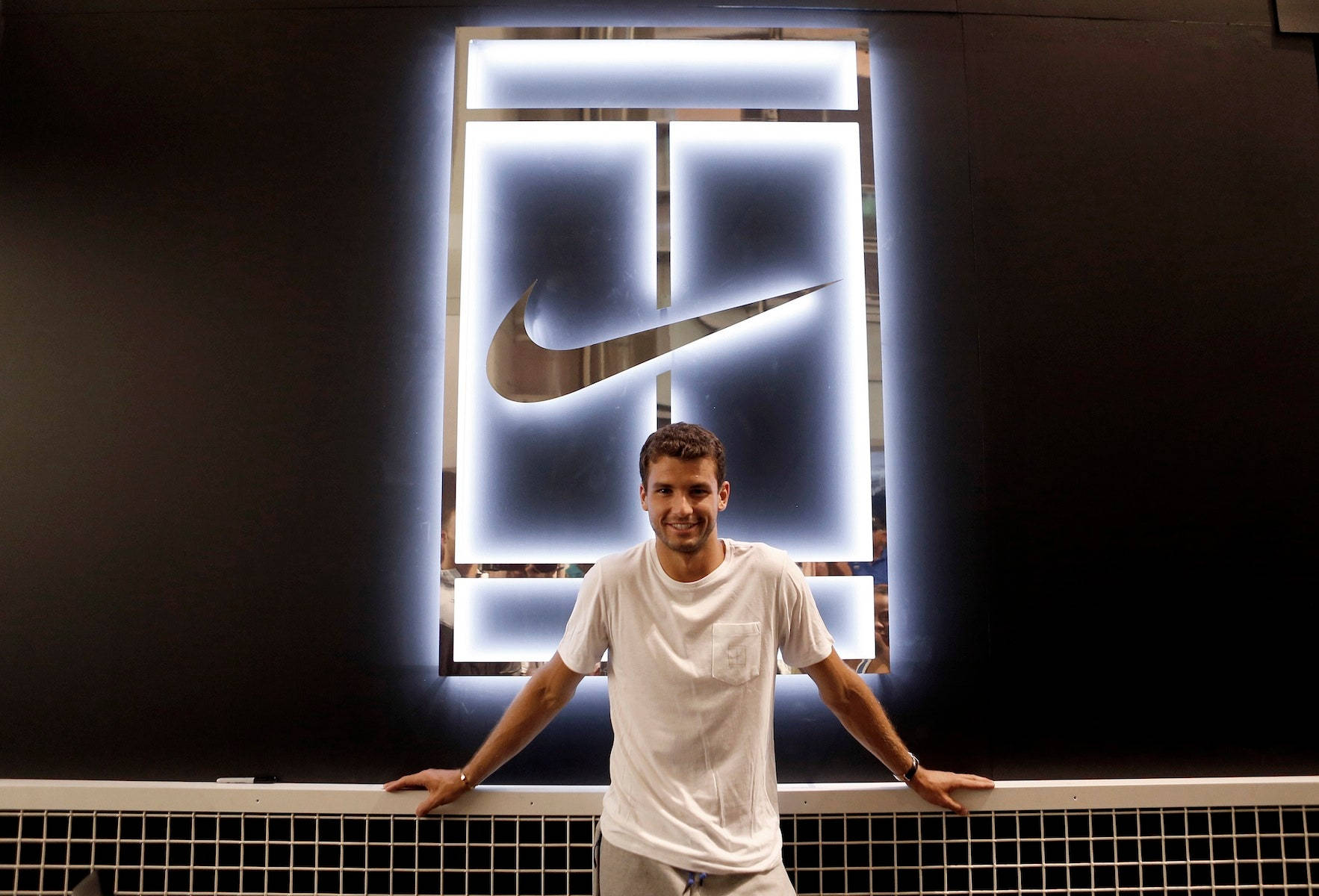 Tennis 桌布 壁紙 — 一個微笑的男人穿著白色T恤，靠在一個發光的Nike標誌牆前的網球網上。