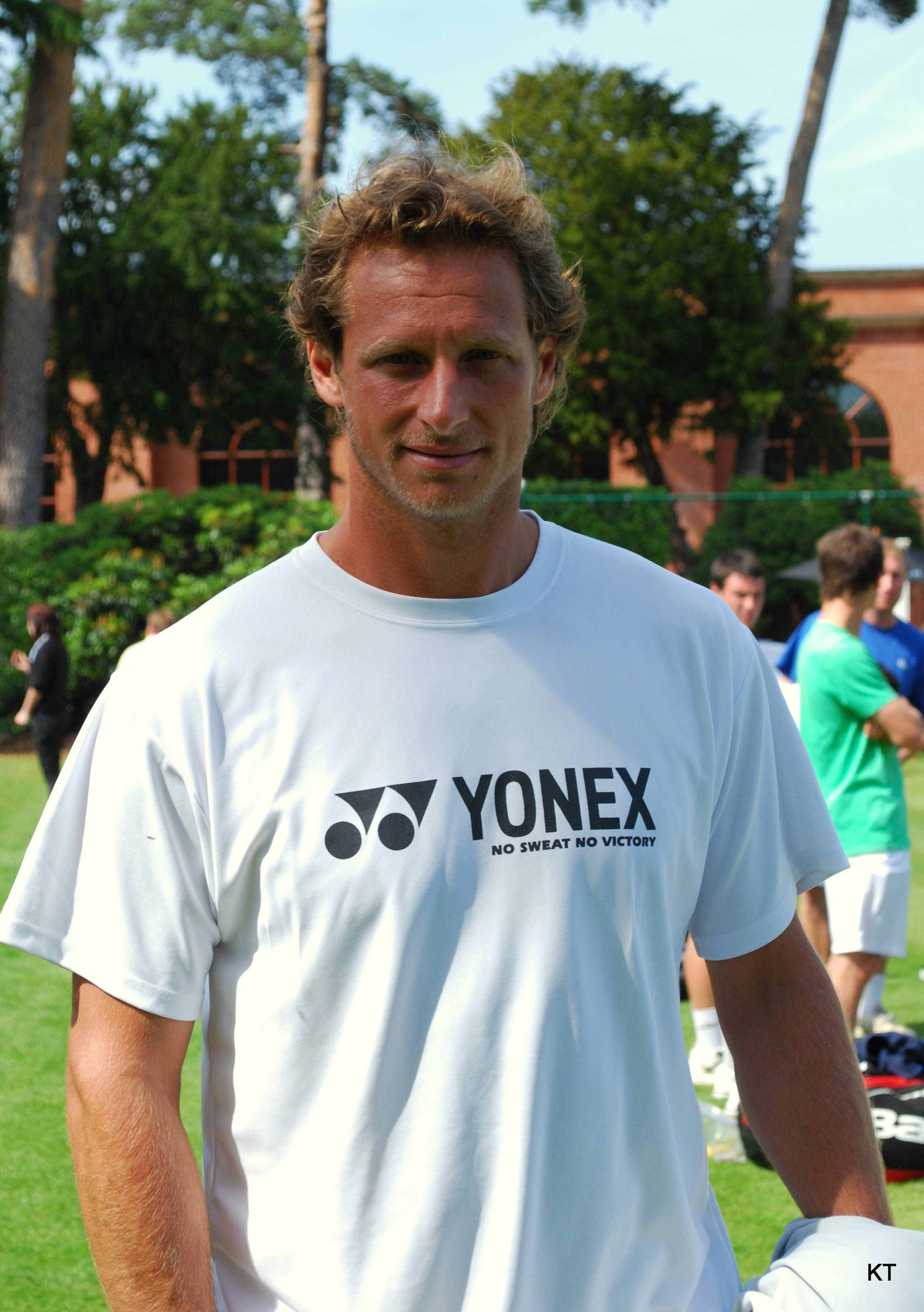 運動 桌布 壁紙 — 一個微笑的男人穿著白色Yonex T恤，在陽光明媚的日子裡站在戶外，身後有樹木和磚砌建築。