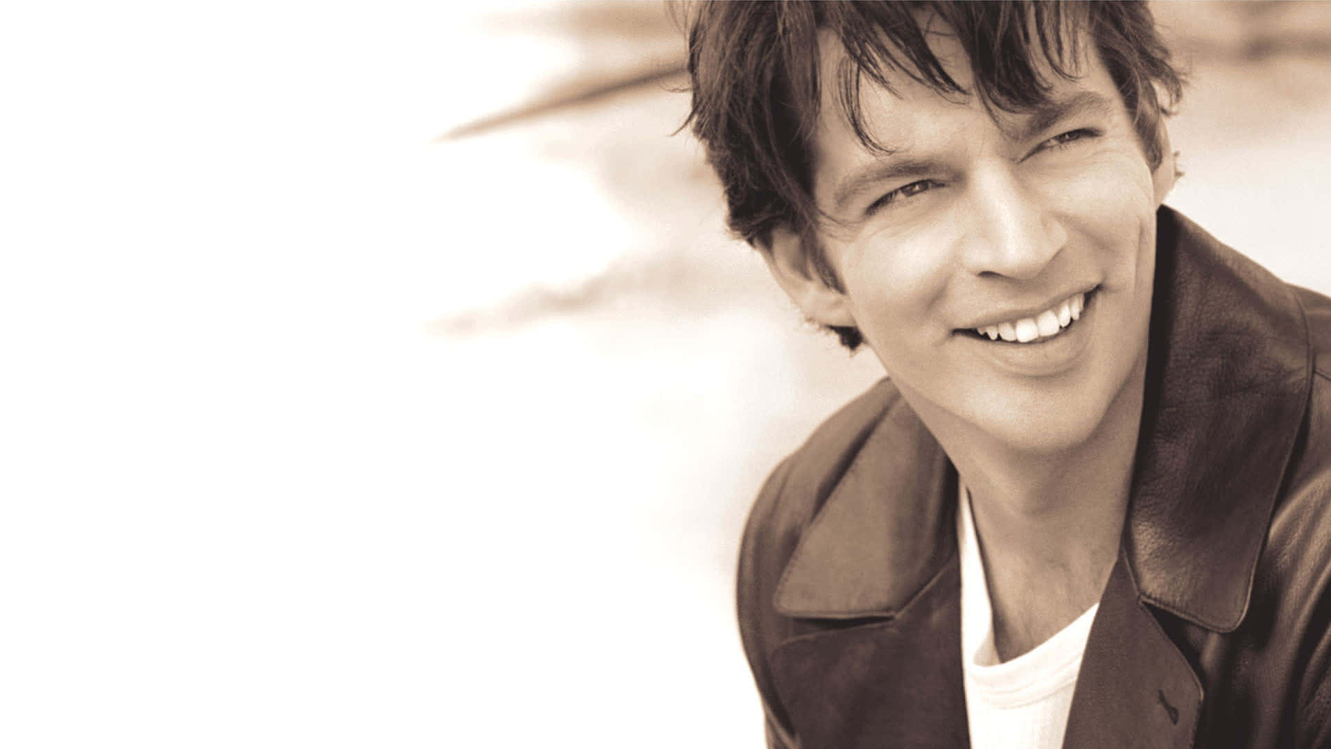 Harry Connick Jr 桌布 壁紙 — 一個微笑的男人穿著皮夾克，在明亮模糊的背景下溫暖地看著鏡頭。