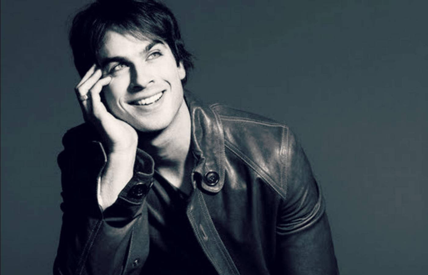 Damon Salvatore 桌布 壁紙 — 一個微笑的男人穿著皮夾克，在柔和的灰色背景上將下巴擱在手上。