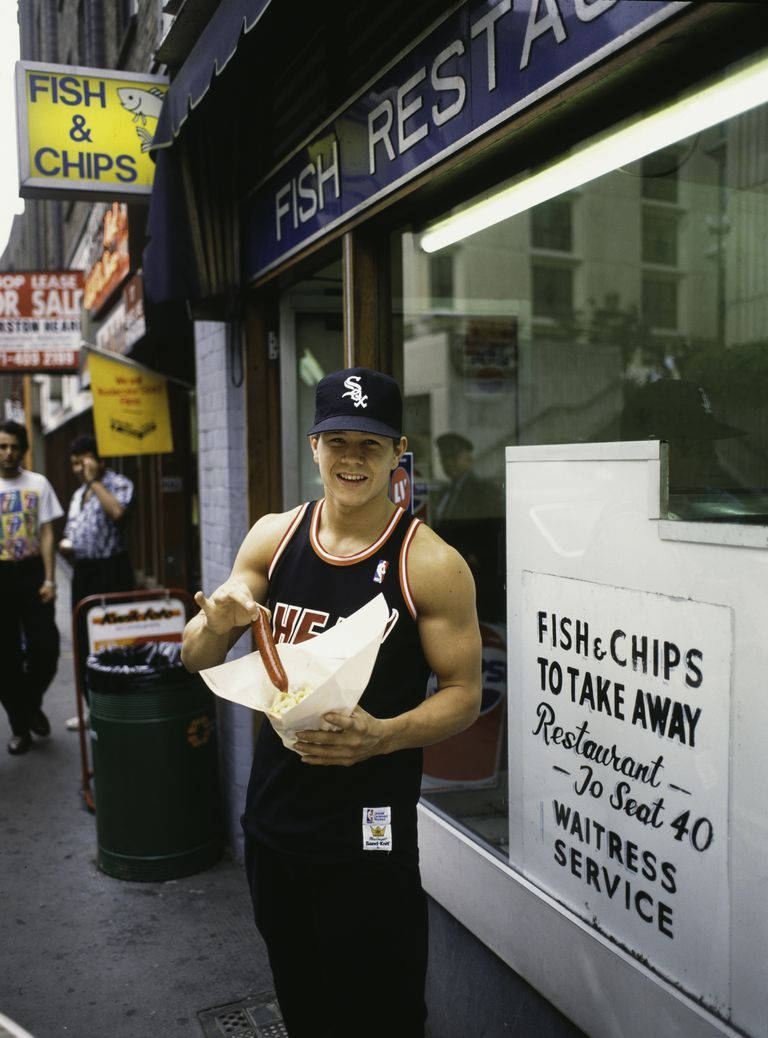Mark Wahlberg 桌布 壁紙 — 一個微笑的男人穿著籃球球衣，在繁忙的街邊餐廳外拿著一個裝有炸魚薯條的紙筒。