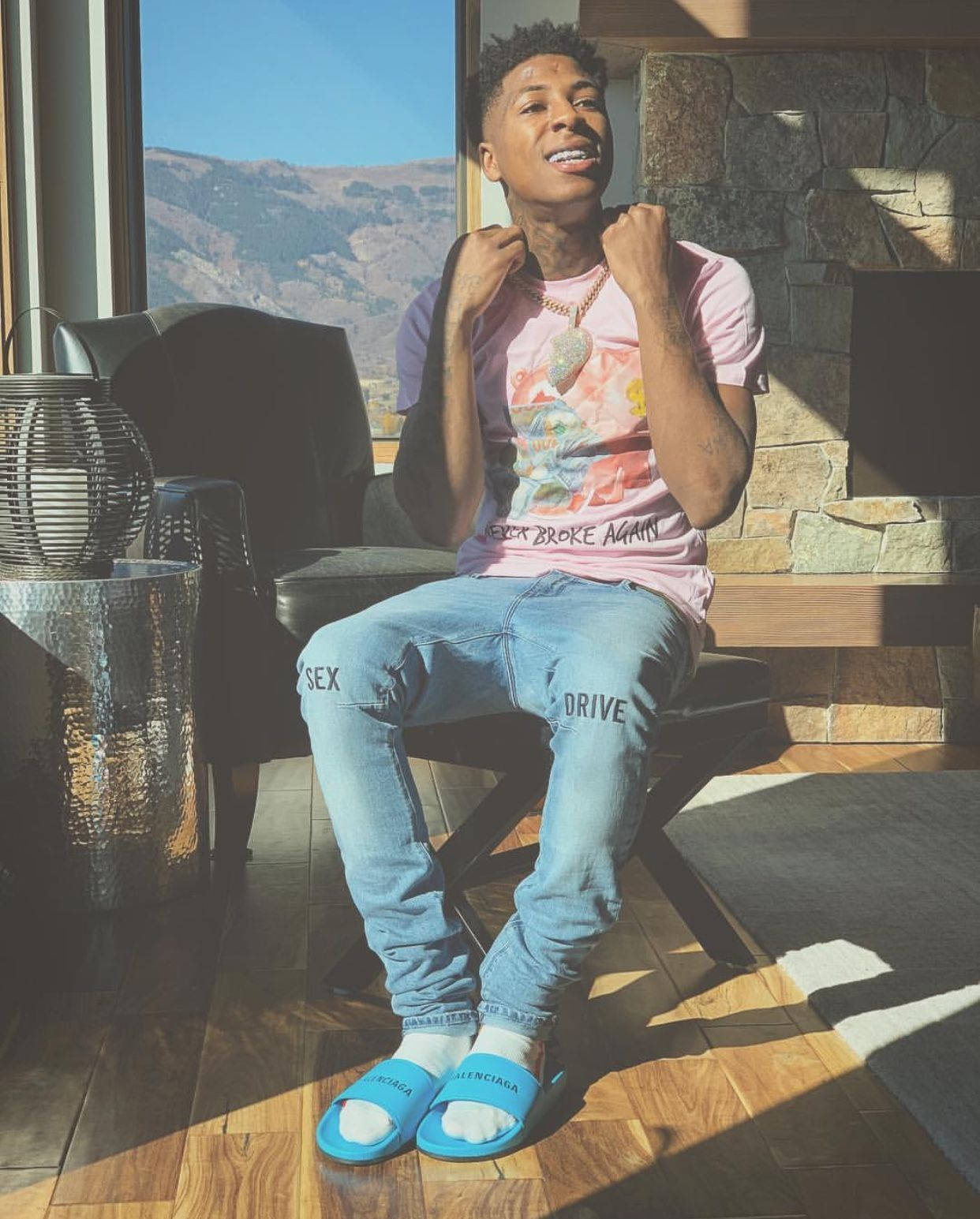 Youngboy 桌布 壁紙 — 一個微笑的男人穿著粉紅色襯衫和藍色牛仔褲，坐在室內窗邊，窗外是沐浴在明亮陽光下的山脈。