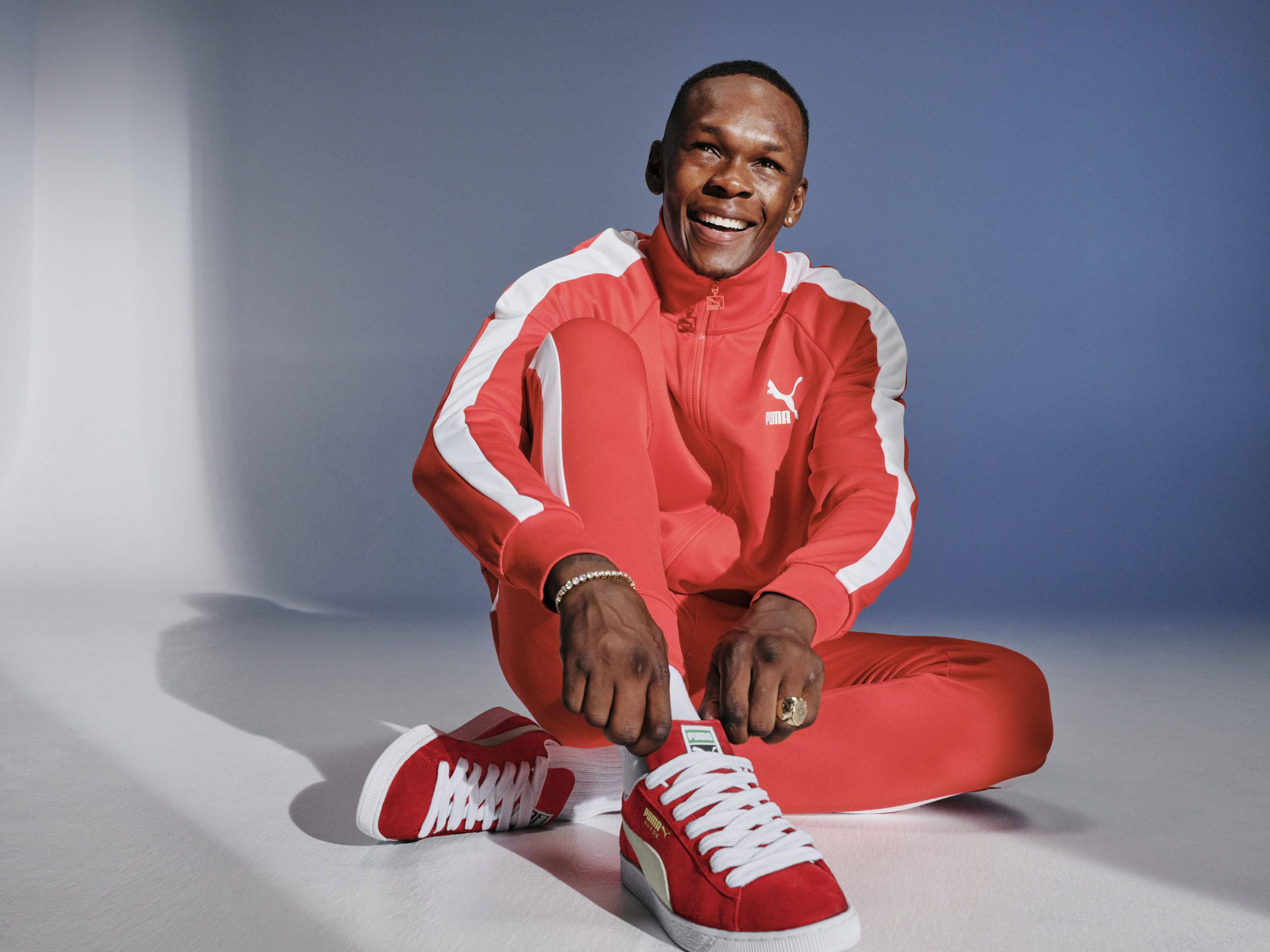 Adesanya 桌布 壁紙 — 一個微笑的男人穿著紅色Puma運動服坐在攝影棚地板上，在漸變藍白背景下繫著他的紅色運動鞋。