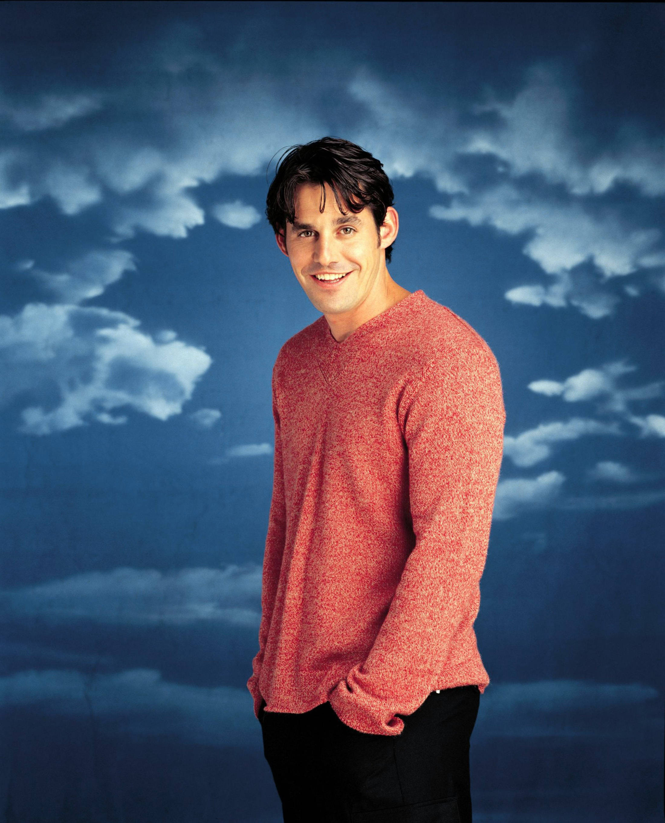 Nicholas Brendon 桌布 壁紙 — 一個微笑的男人穿著紅色毛衣，自信地站在戲劇性的多雲藍天背景前。