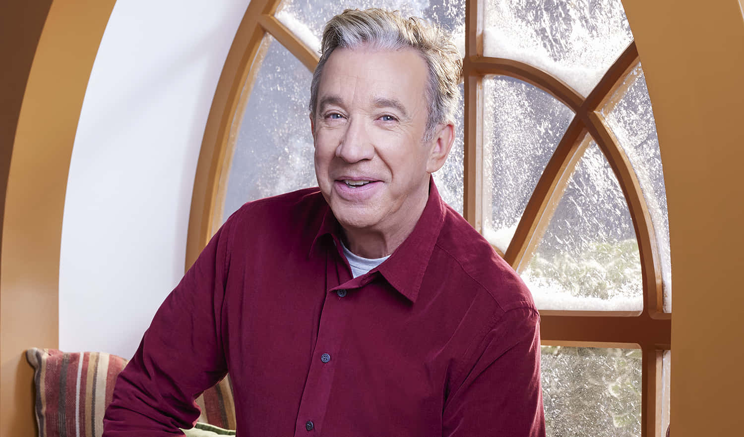 Tim Allen 桌布 壁紙 — 一個微笑的男人穿著紅色襯衫，坐在一個有磨砂玻璃板的大拱形窗旁邊。