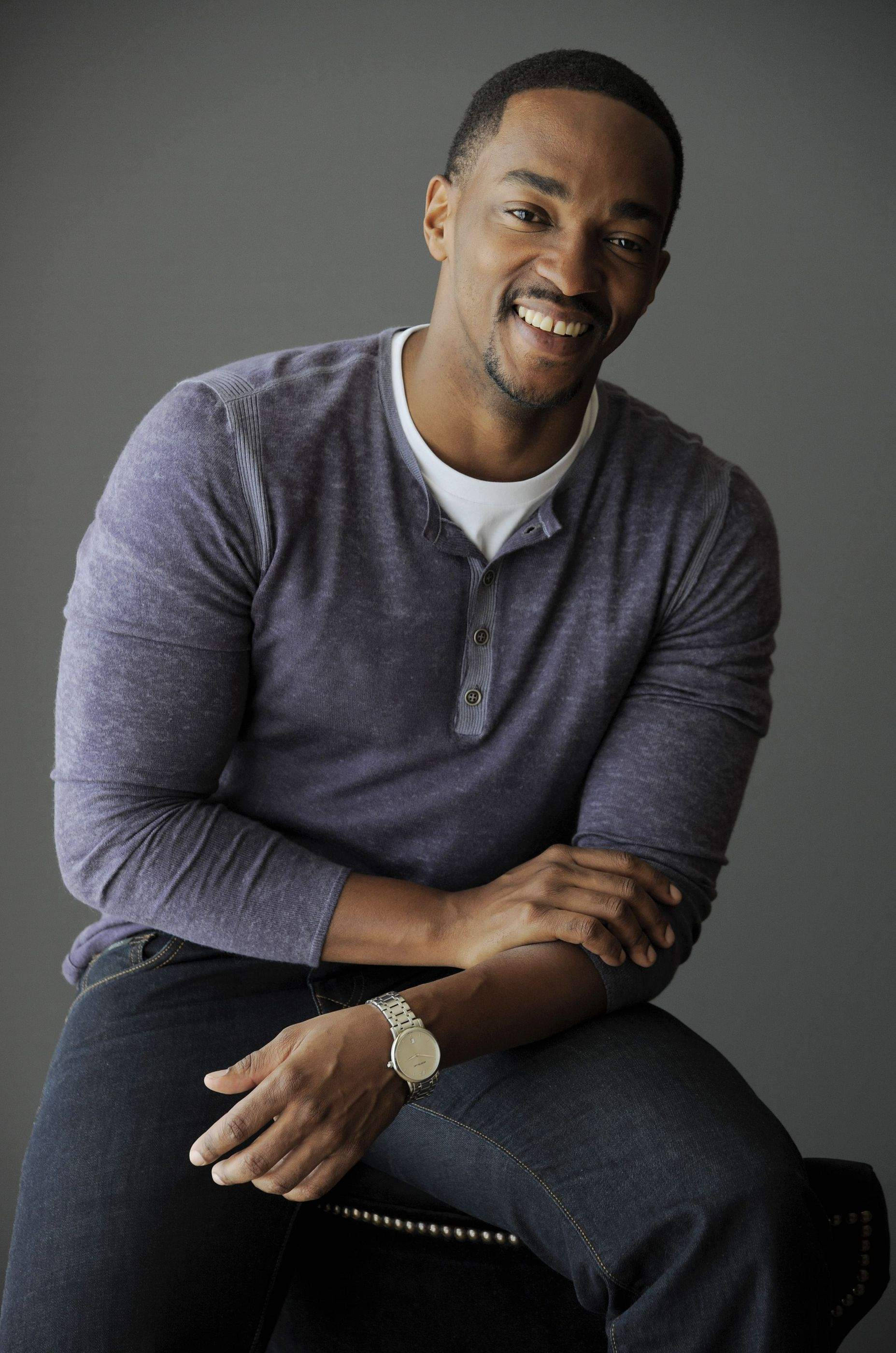 Anthony Mackie 桌布 壁紙 — 一個微笑的男人穿著紫色亨利衫，隨意地坐在椅子上，背景是純灰色。