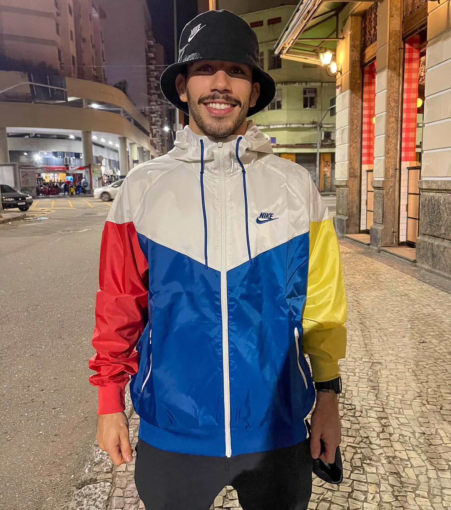 Celebrity 桌布 壁紙 — 一個微笑的男人穿著色彩繽紛的Nike風衣和漁夫帽，在夜晚的鵝卵石街道上，站在溫暖的路燈下。