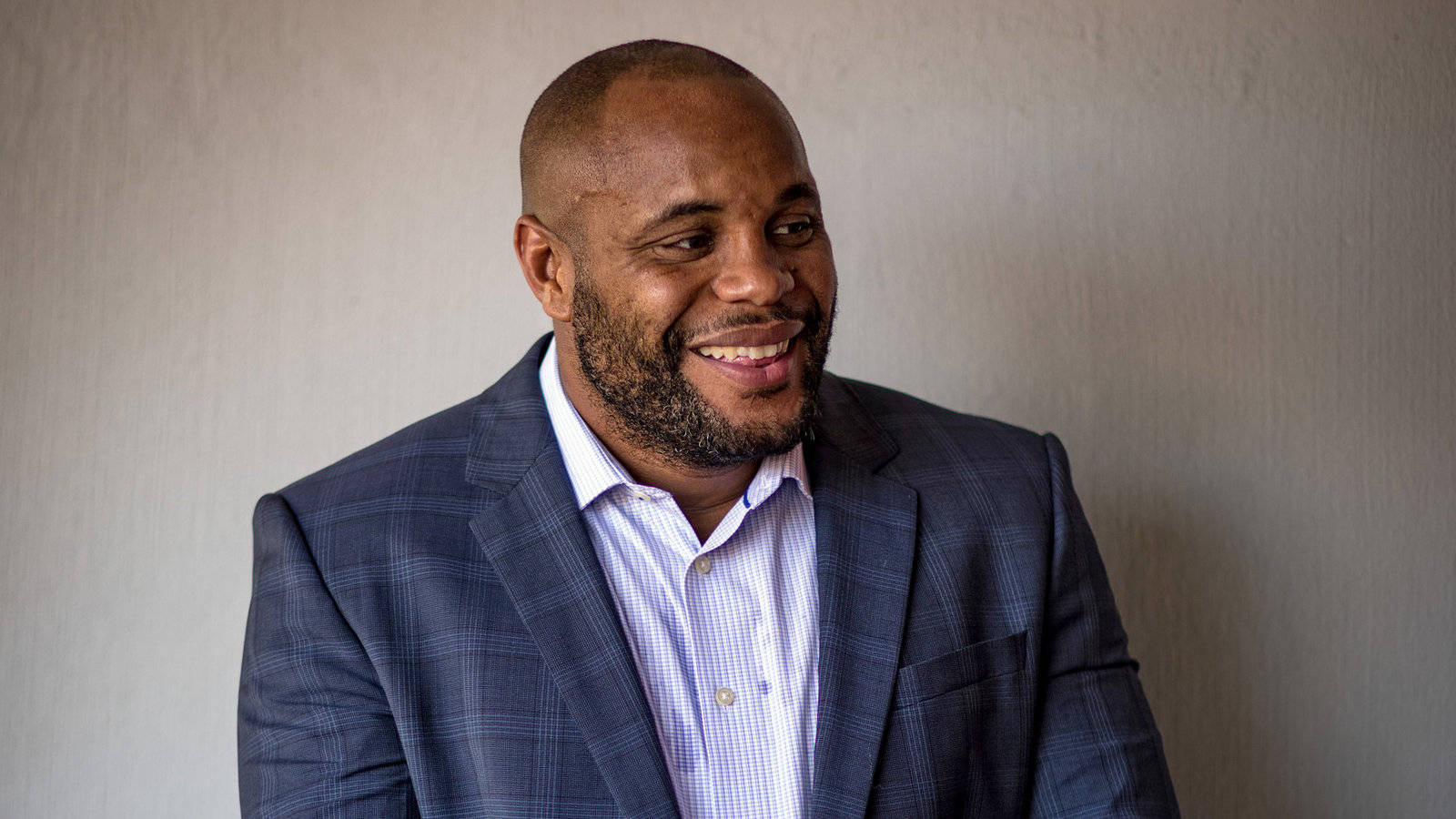 UFC 桌布 壁紙 — 一個微笑的男人穿著藍色格紋西裝和條紋襯衫，站在樸素的紋理牆前。