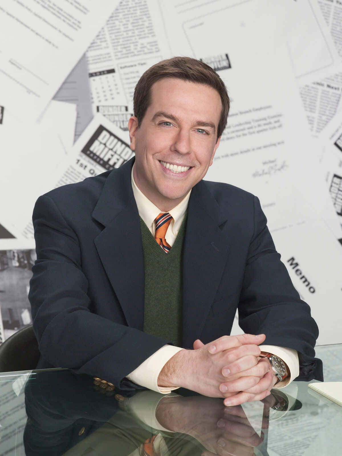 ed helms 桌布 壁紙 — 一個微笑的男人穿著西裝打領帶，坐在玻璃桌前，身後散落著文件和備忘錄。