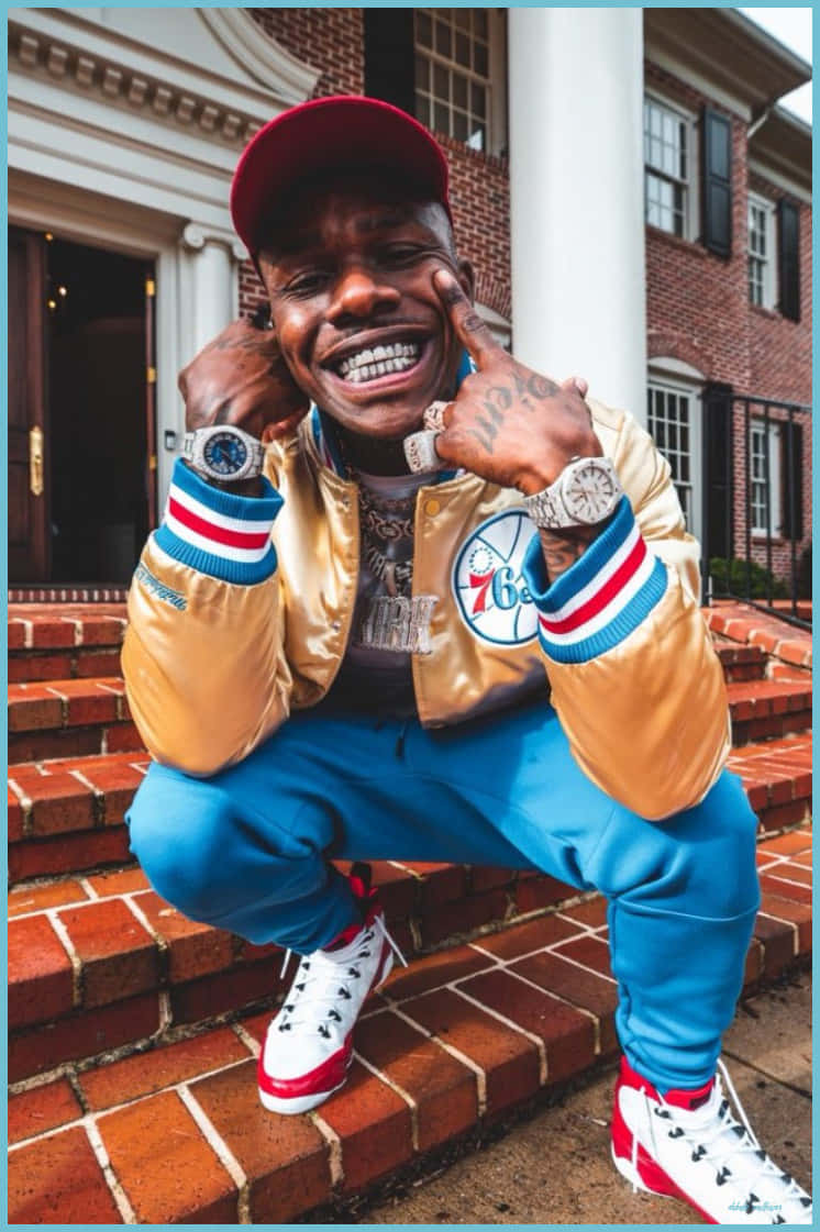 Dababy Cartoon 桌布 壁紙 — 一個微笑的男人穿著金色外套和藍色褲子，蹲在豪宅前的磚階上，展示他的奢華手錶和鑽石牙齒。