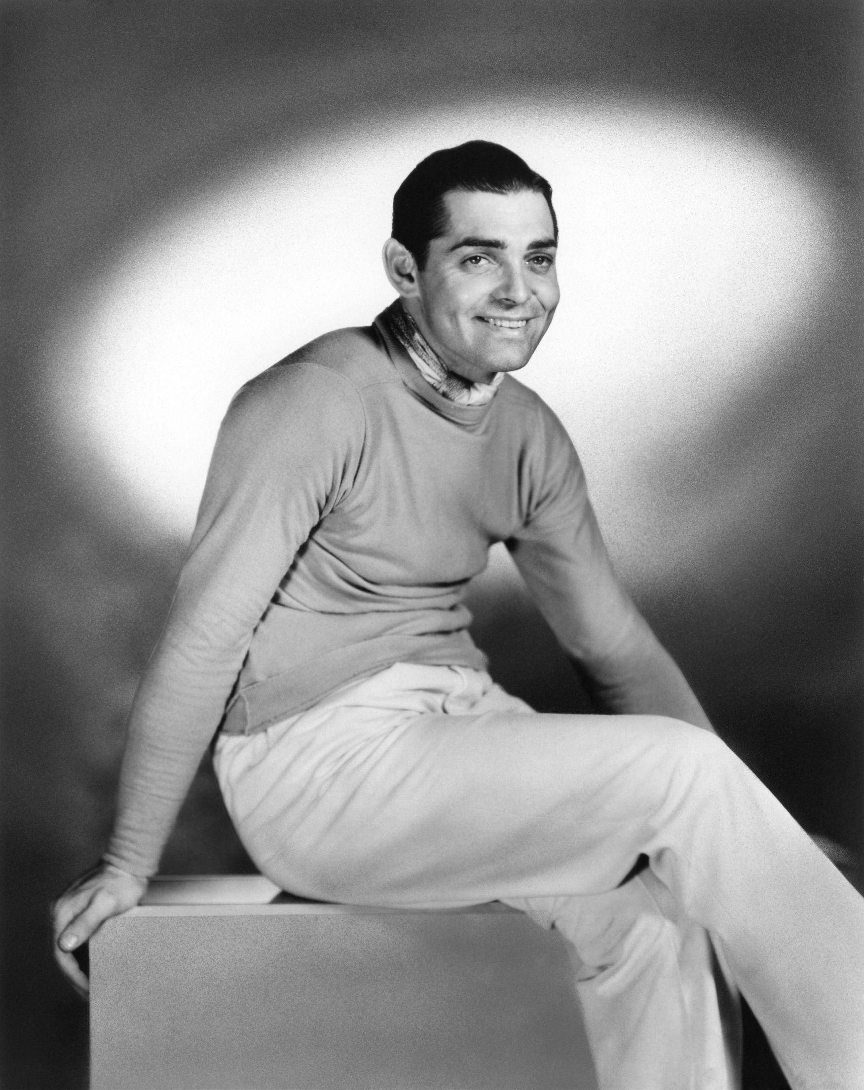 actor 桌布 壁紙 — 一個微笑的男人穿著高領毛衣和淺色褲子，隨意地坐在一個方塊上，背景是柔和照明的攝影棚。