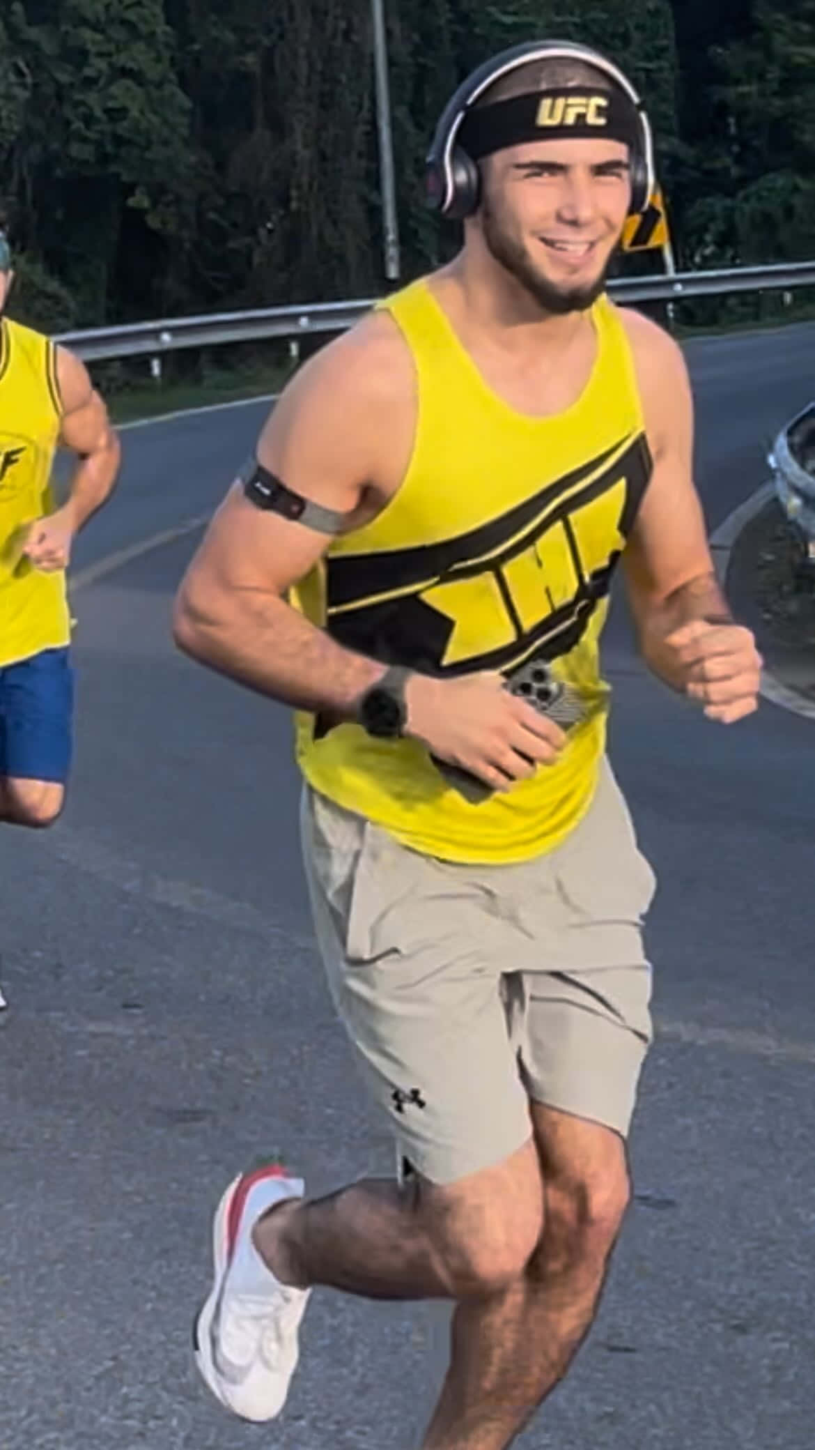 Fitness 桌布 壁紙 — 一個微笑的男人穿著黃色UFC背心和耳機，在鋪砌的道路上跑步，背景有樹木。