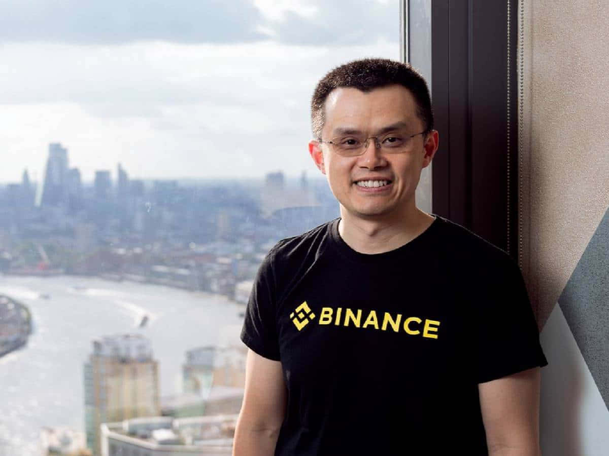 Binance 桌布 壁紙 — 一個微笑的男人穿著黑色Binance T恤，靠著窗戶，窗外是廣闊的城市景觀和一條河流。