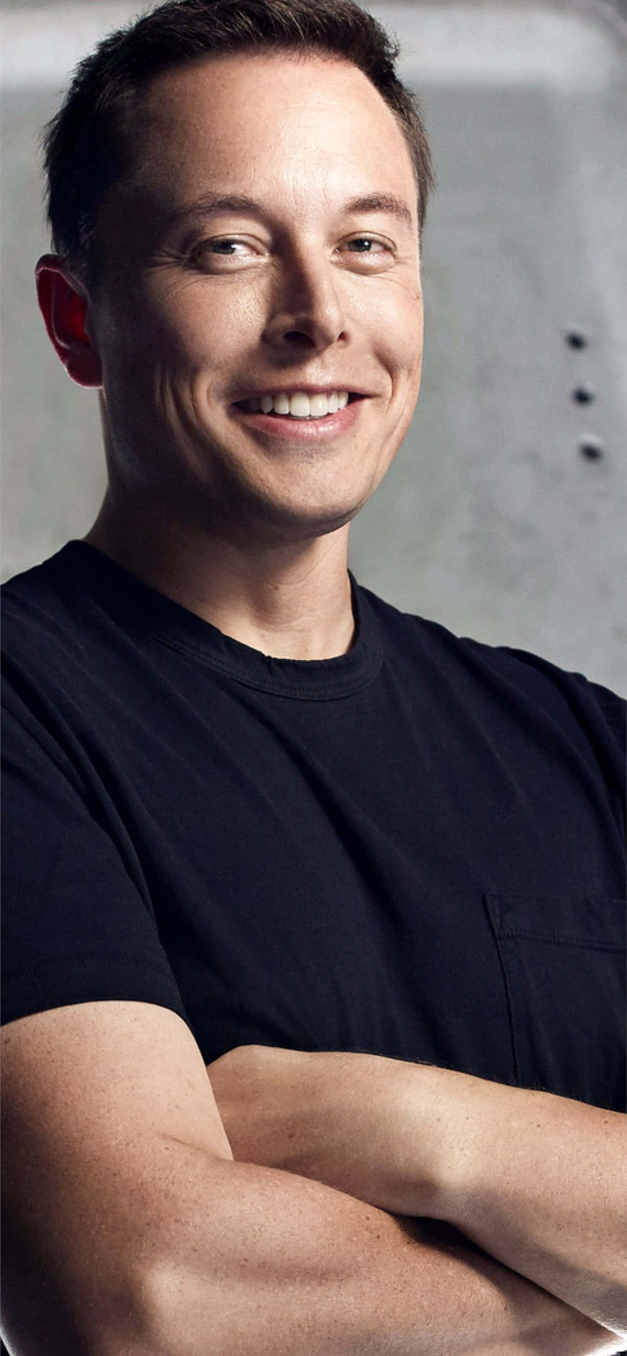 攝影 桌布 壁紙 — 一個微笑的男人穿著黑色T恤，雙臂交叉，自信地站在有紋理的灰色牆前。