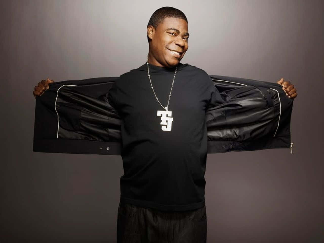 Tracy Morgan 桌布 壁紙 — 一個微笑的男人穿著黑色T恤和鏈條，在純灰色工作室背景前，將黑色外套敞開。