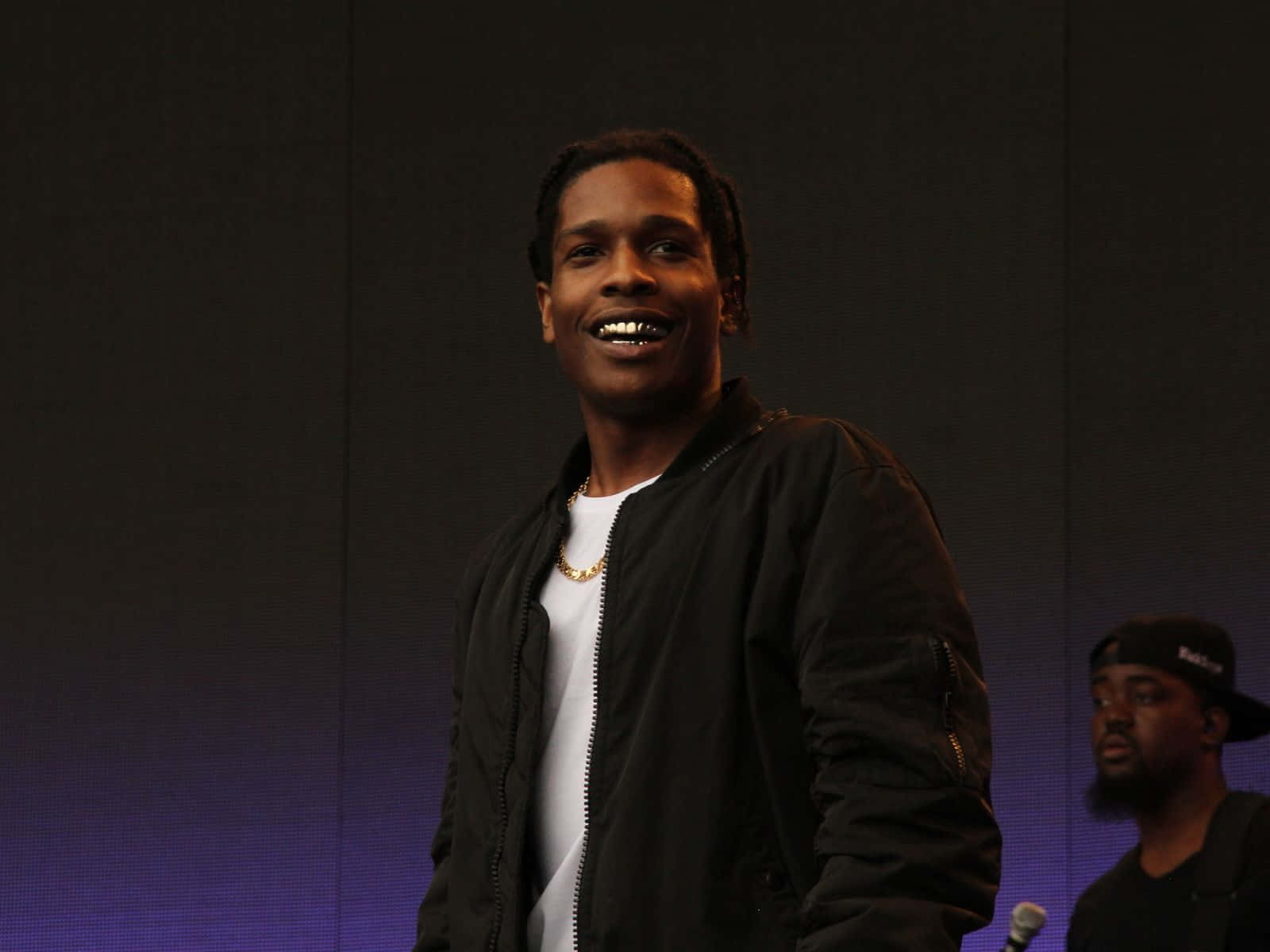 Asap Rocky 桌布 壁紙 — 一個微笑的男人穿著黑色外套和白色T恤，站在一個背後有紫色燈光的舞台上。