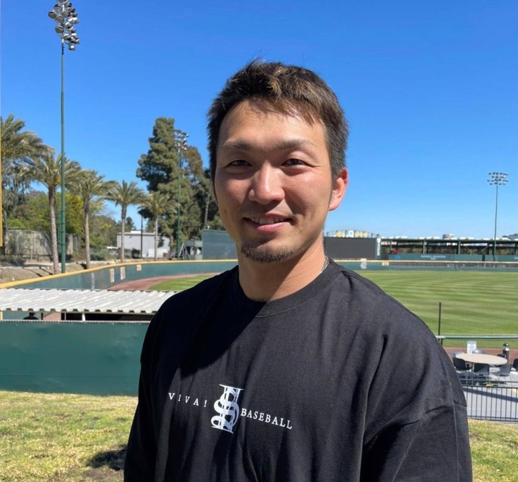 Athlete 桌布 壁紙 — 一個微笑的男人穿著黑色棒球T恤，站在陽光明媚的棒球場上，有棕櫚樹和體育場燈光，背景是晴朗的藍天。