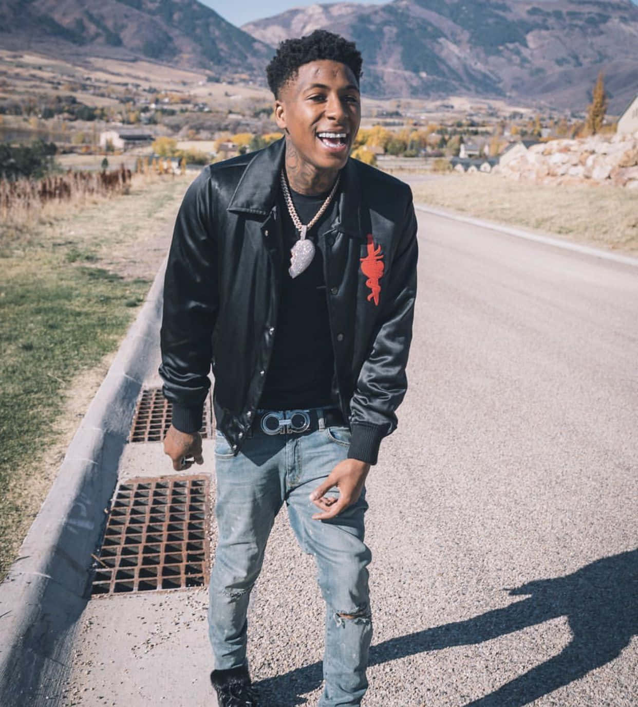 Youngboy 桌布 壁紙 — 一個微笑的男人穿著黑色皮夾克和破洞牛仔褲，站在路邊，背景有山脈和秋天的樹木。