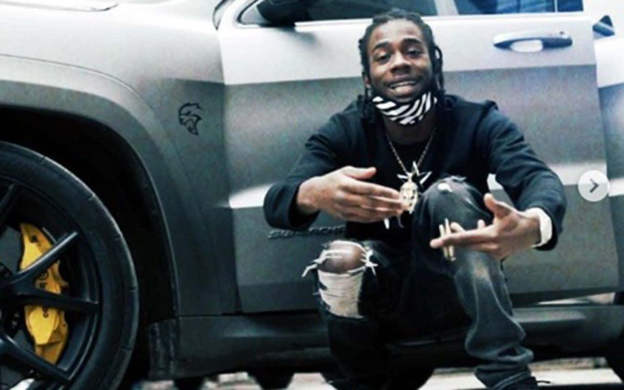 Dead Rappers 桌布 壁紙 — 一個微笑的男人穿著黑色破洞牛仔褲和連帽衫，蹲在一輛帶有黃色制動卡鉗的磨砂灰色SUV旁邊。