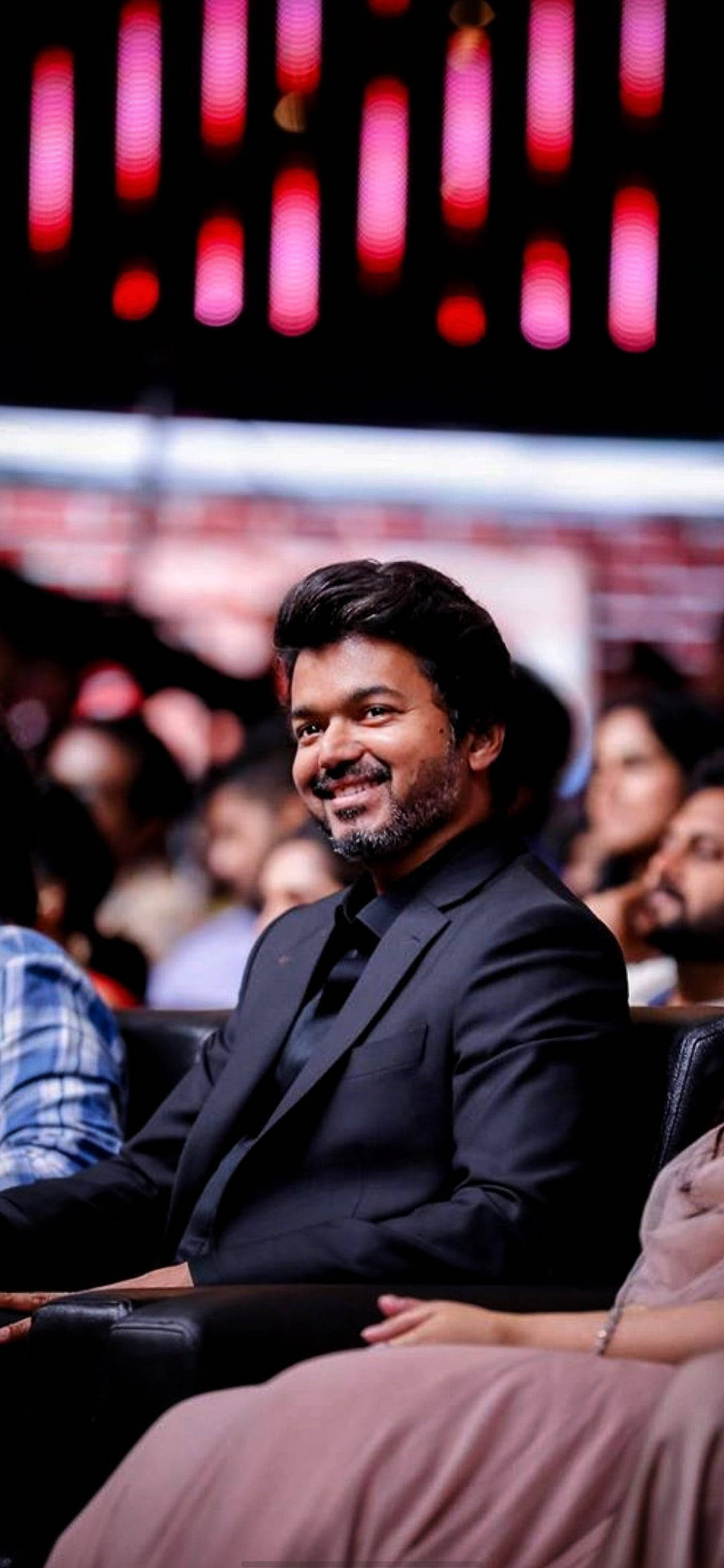 vijay HD 桌布 壁紙 — 一個微笑的男人穿著黑色西裝坐在燈光昏暗的禮堂中，背景有發光的紅色垂直燈光。