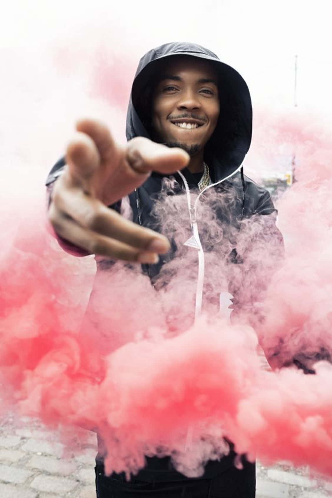 Herbo iPhone 桌布 壁紙 — 一個微笑的男人穿著黑色連帽衫，在城市的街道上伸手穿過充滿活力的粉紅色煙霧。